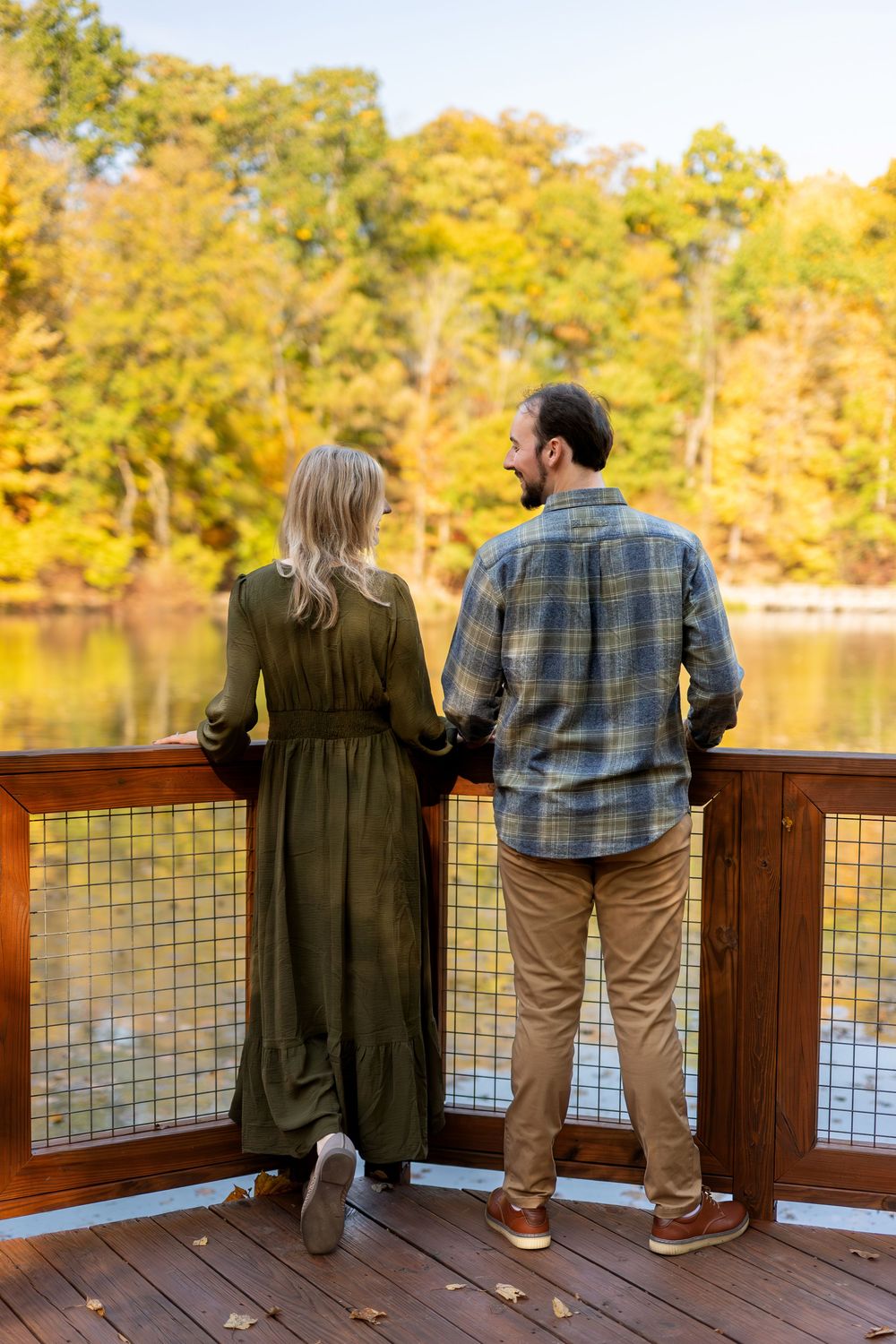 Couple holding hands while gazing at a lake surrounded by colorful autumn foliage from a deck.