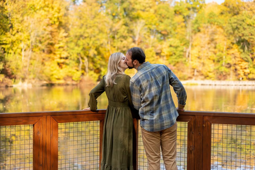 Couple sharing a romantic kiss on a wooden deck overlooking a lake with fall foliage.