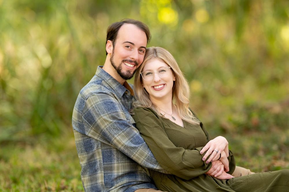 Smiling couple sitting closely together outdoors in a lush green autumn setting.