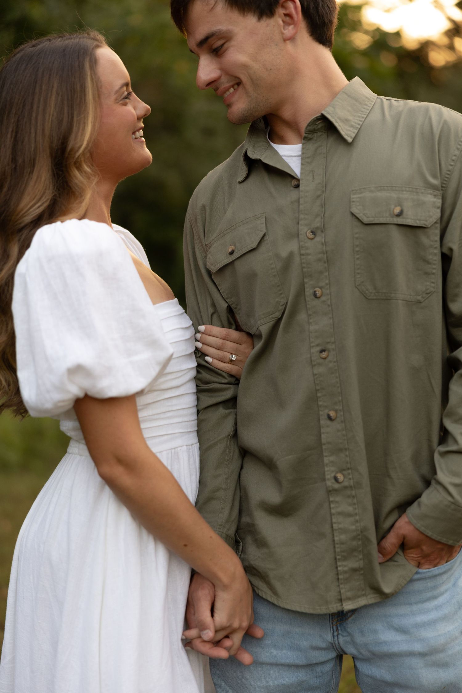 A couple shares a romantic moment while holding hands in a park with sunlit trees in the background.