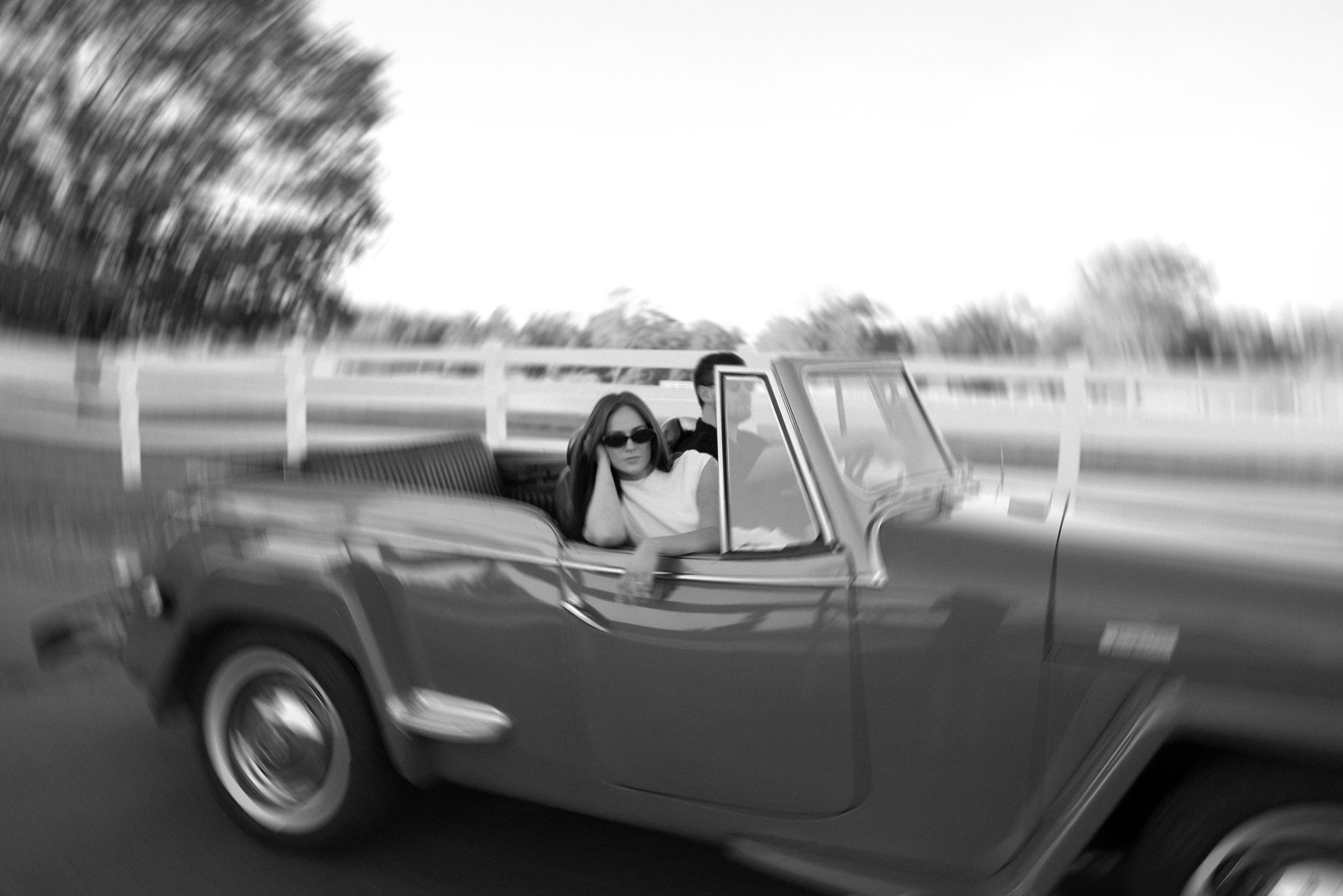 A black and white motion blur photo of newlyweds riding in a vintage convertible car.