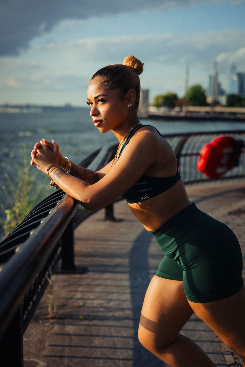 Athletic woman in workout attire stretches on waterfront boardwalk during sunset with city skyline in background.