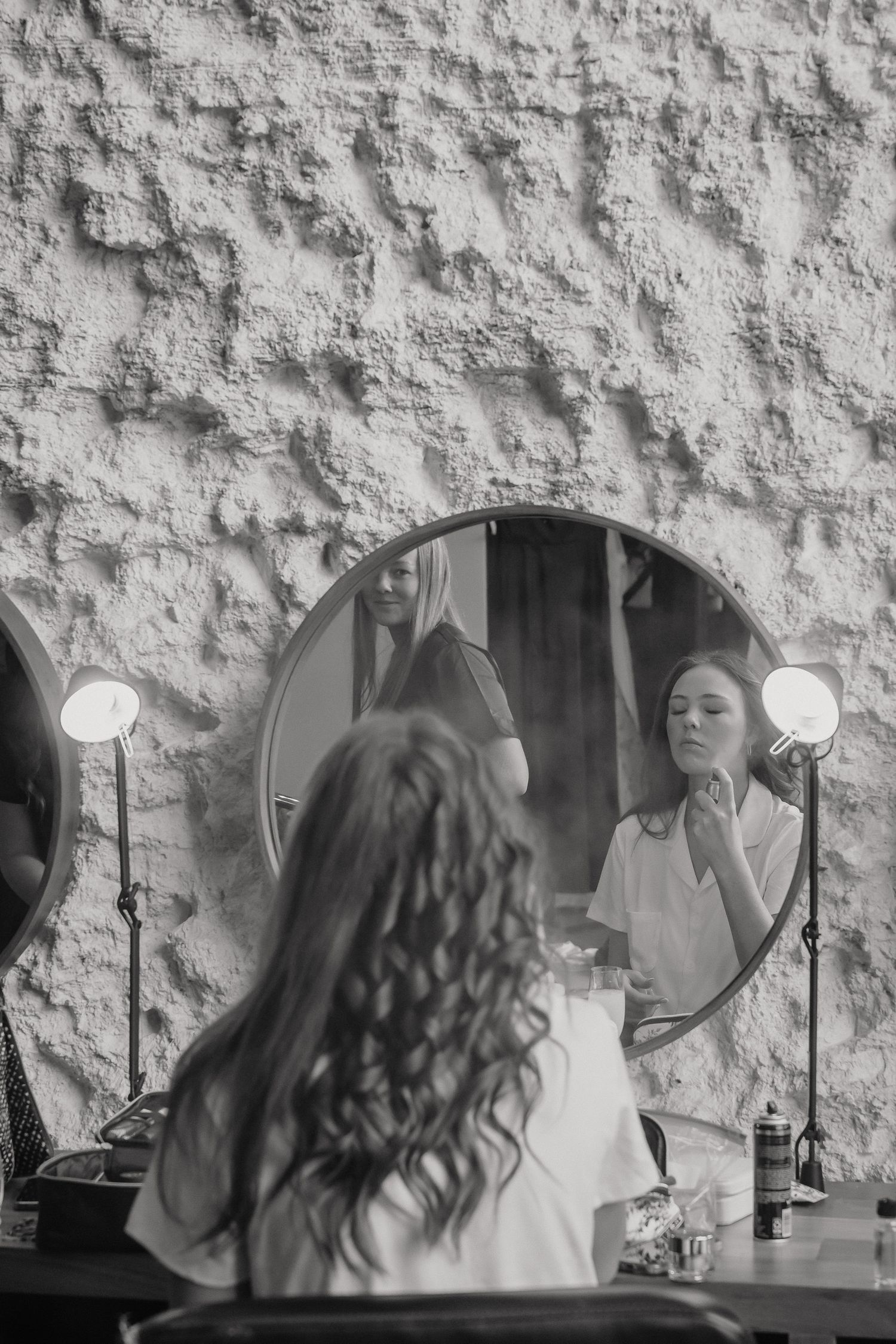 Black and white photograph of a bridal preparation scene with soft lighting and textured wall backdrop.