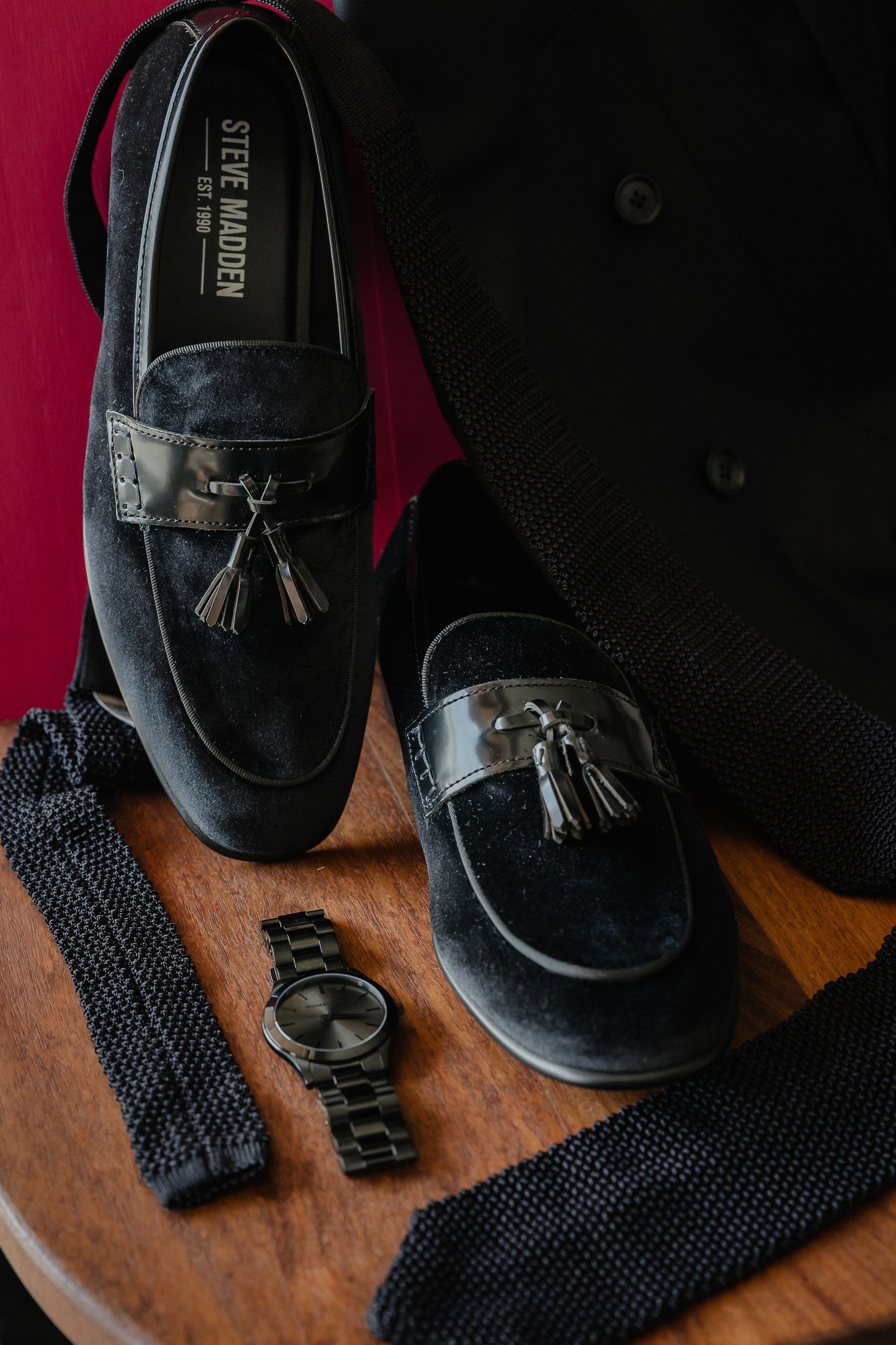 Black leather dress shoes with tassels arranged on a brown leather surface with dark fabric accessories.
