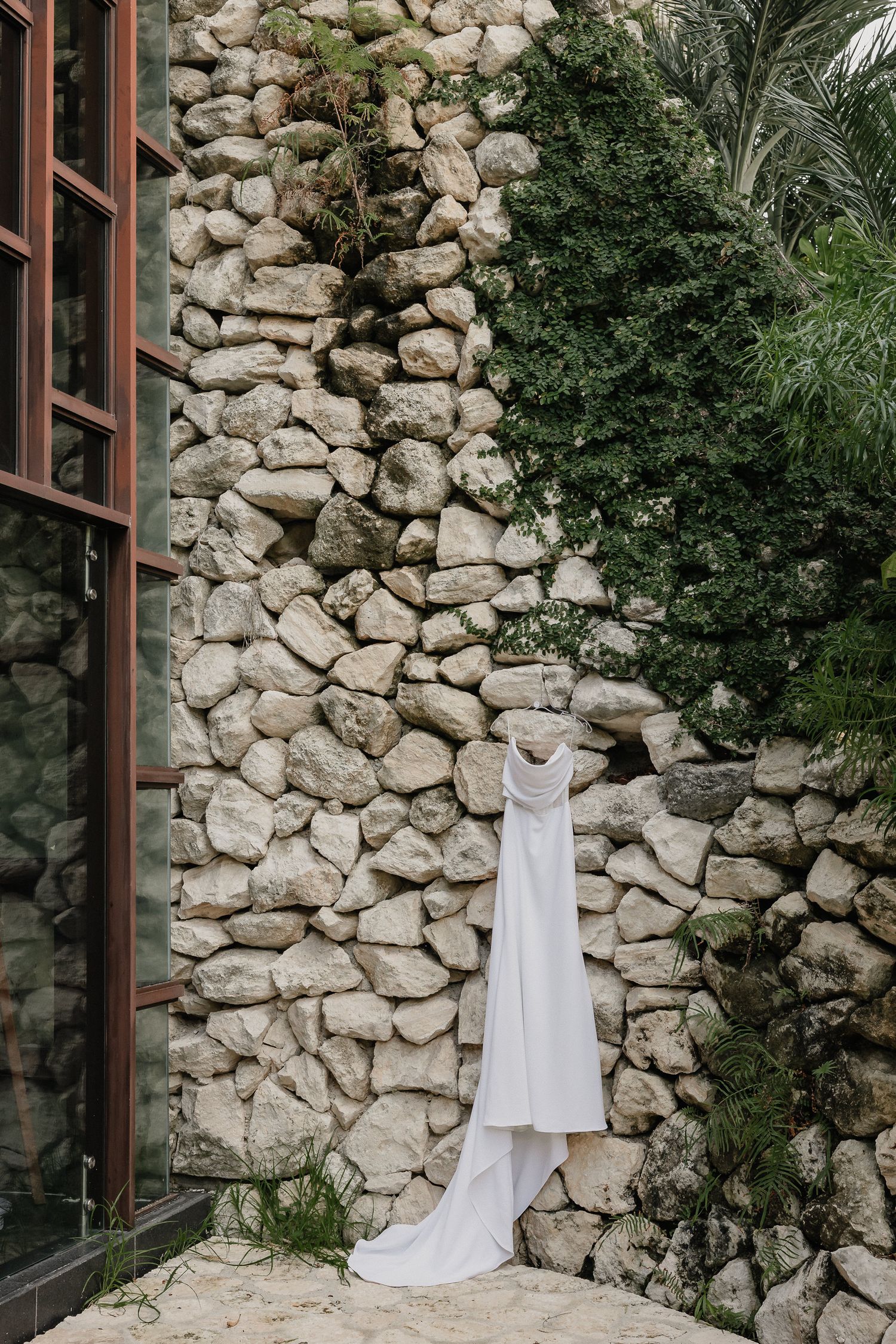 White wedding dress displayed against rustic stone wall with natural greenery.
