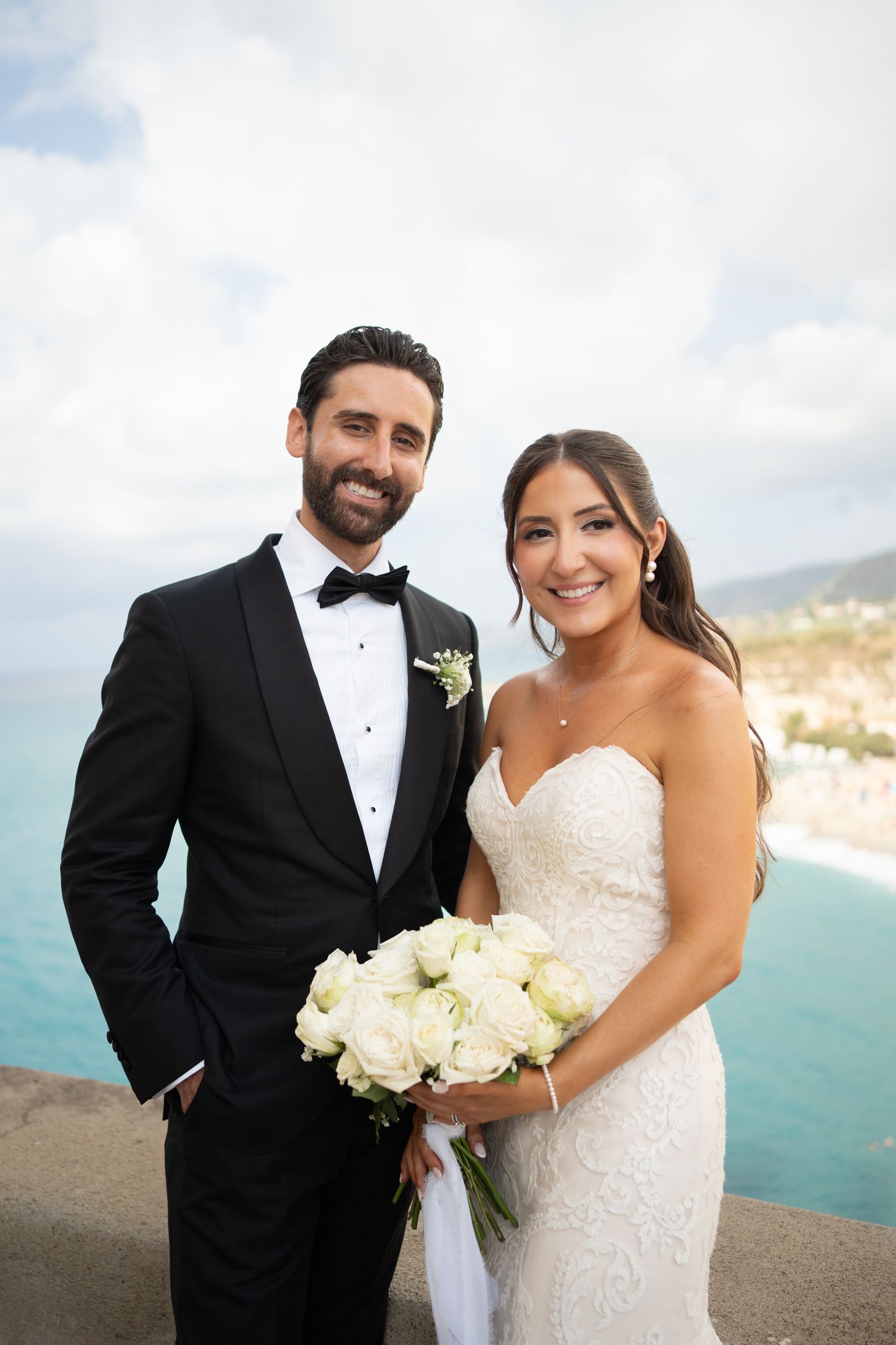 A newlywed couple poses for wedding photos on a scenic coastal cliff overlooking a beautiful blue ocean bay.