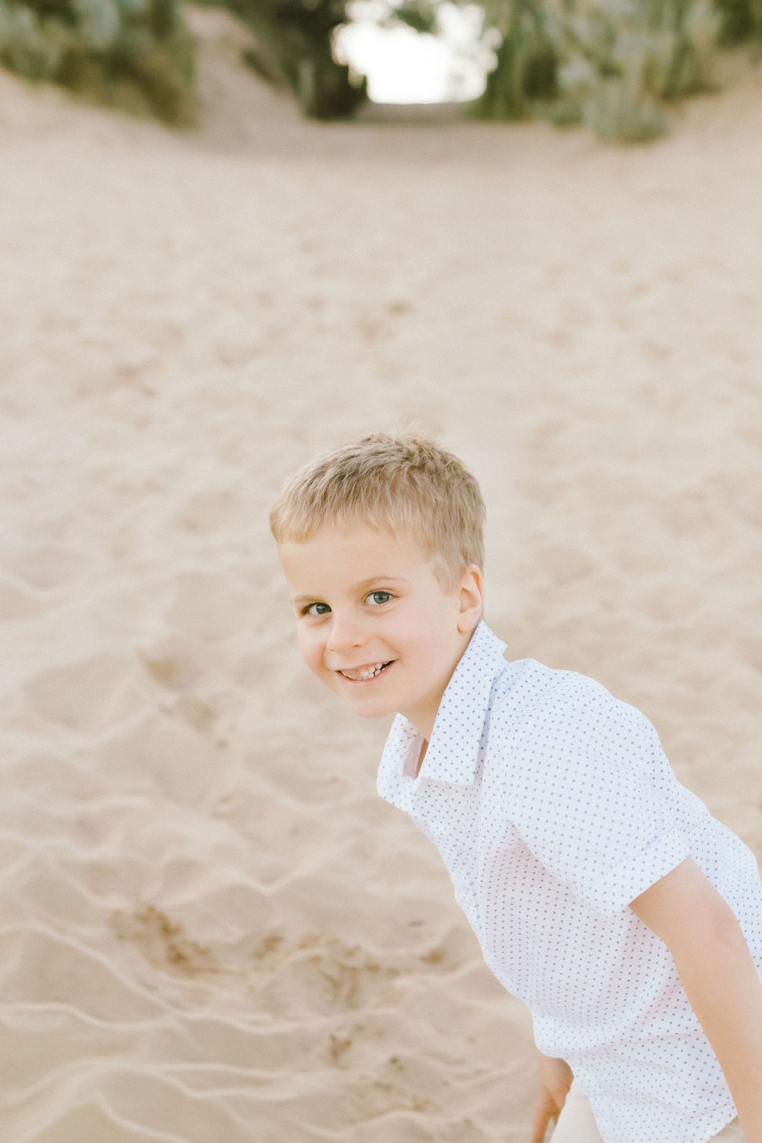 A child in a white checkered shirt plays and smiles while sitting in soft beach sand on a sunny day.