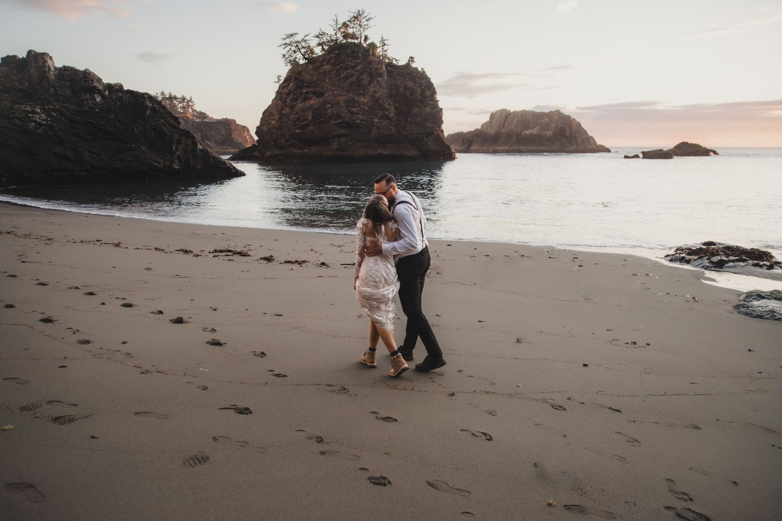 Couple embraces on a scenic beach with rocky cliffs and ocean waves during golden hour sunset.