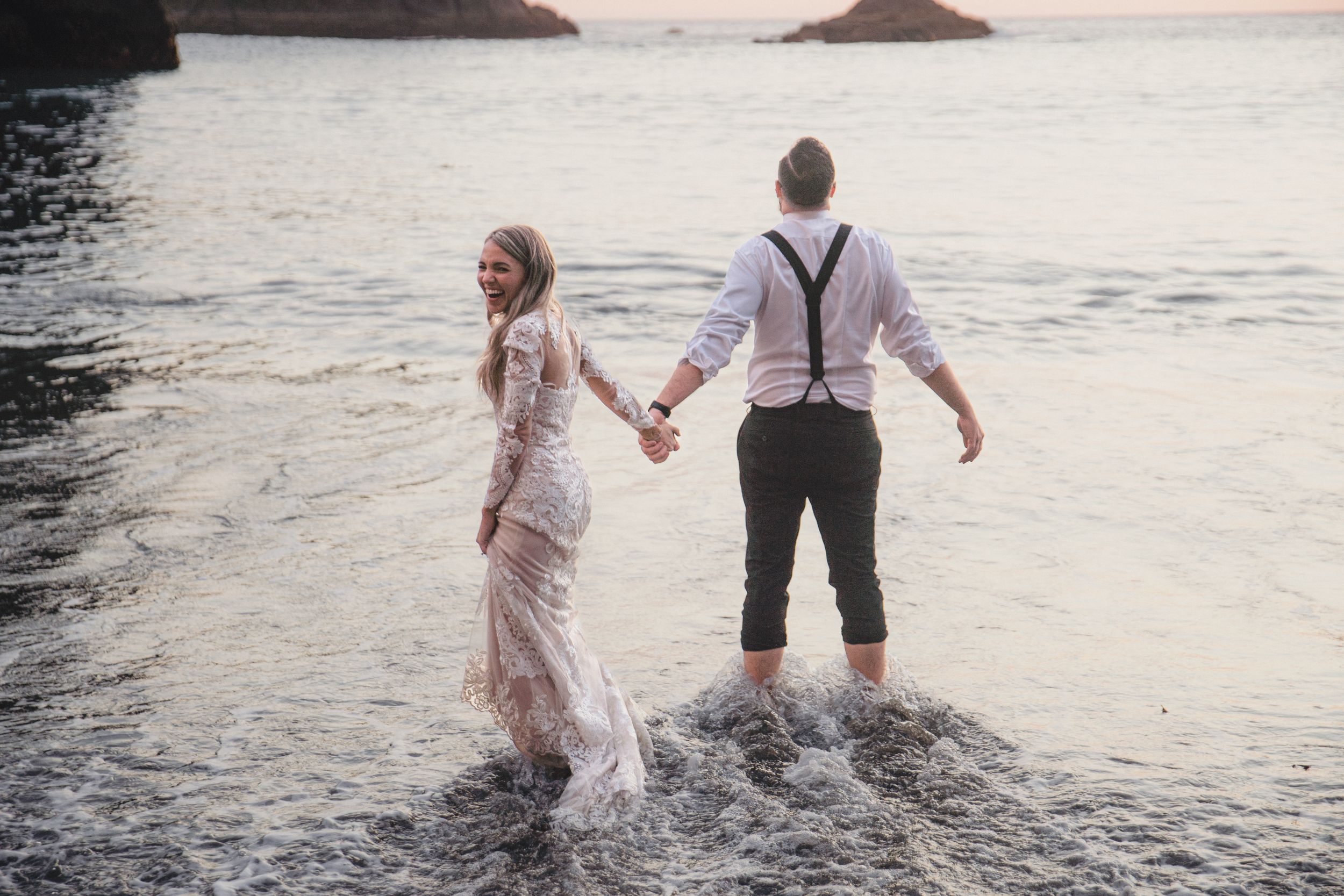 Couple splashing in ocean waves at sunset near rugged coastal rocks.