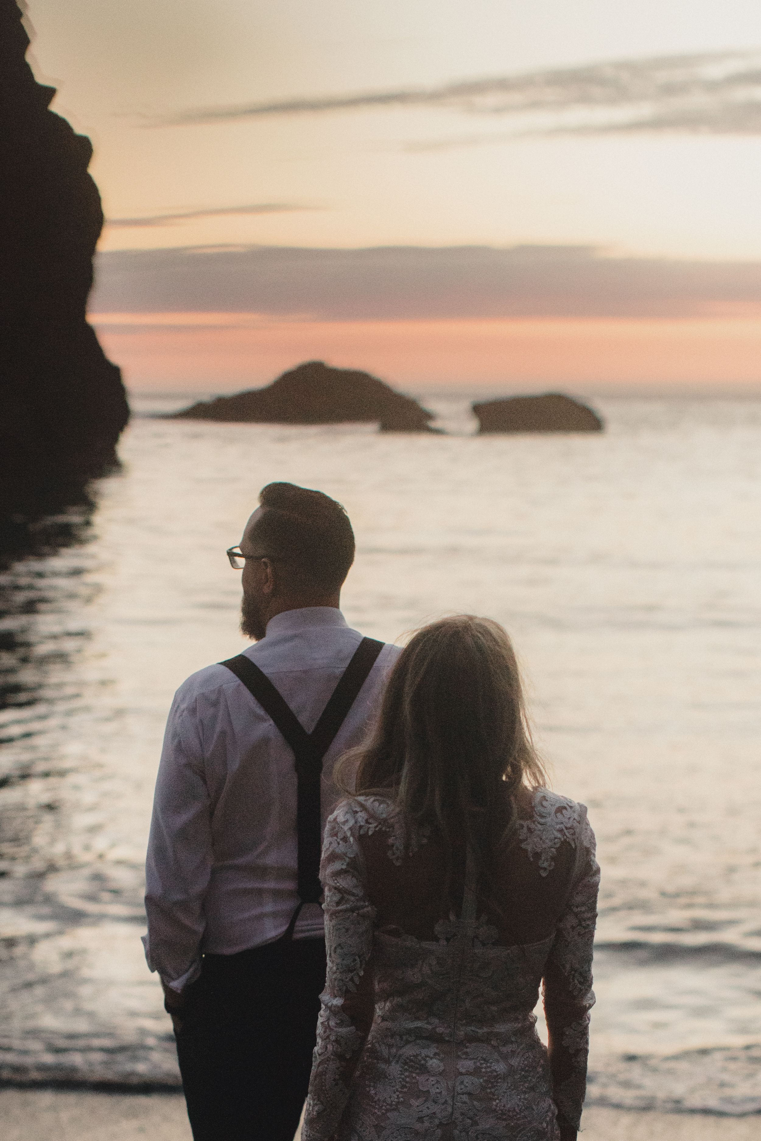Two figures watching the sunset over the Pacific Ocean from a rocky beach overlook.