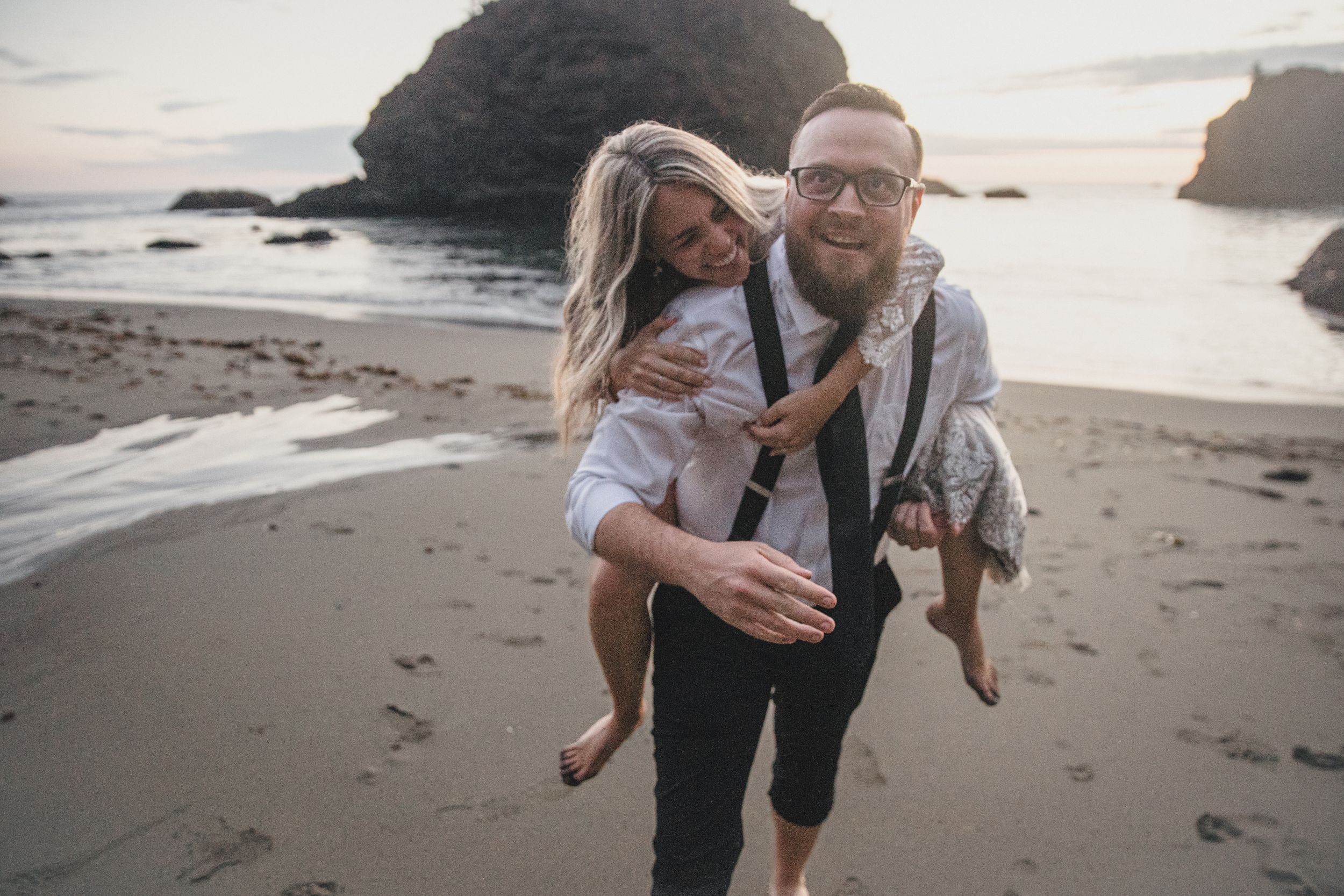 Couple playing and laughing together on a misty beach with rocky cliffs in background.