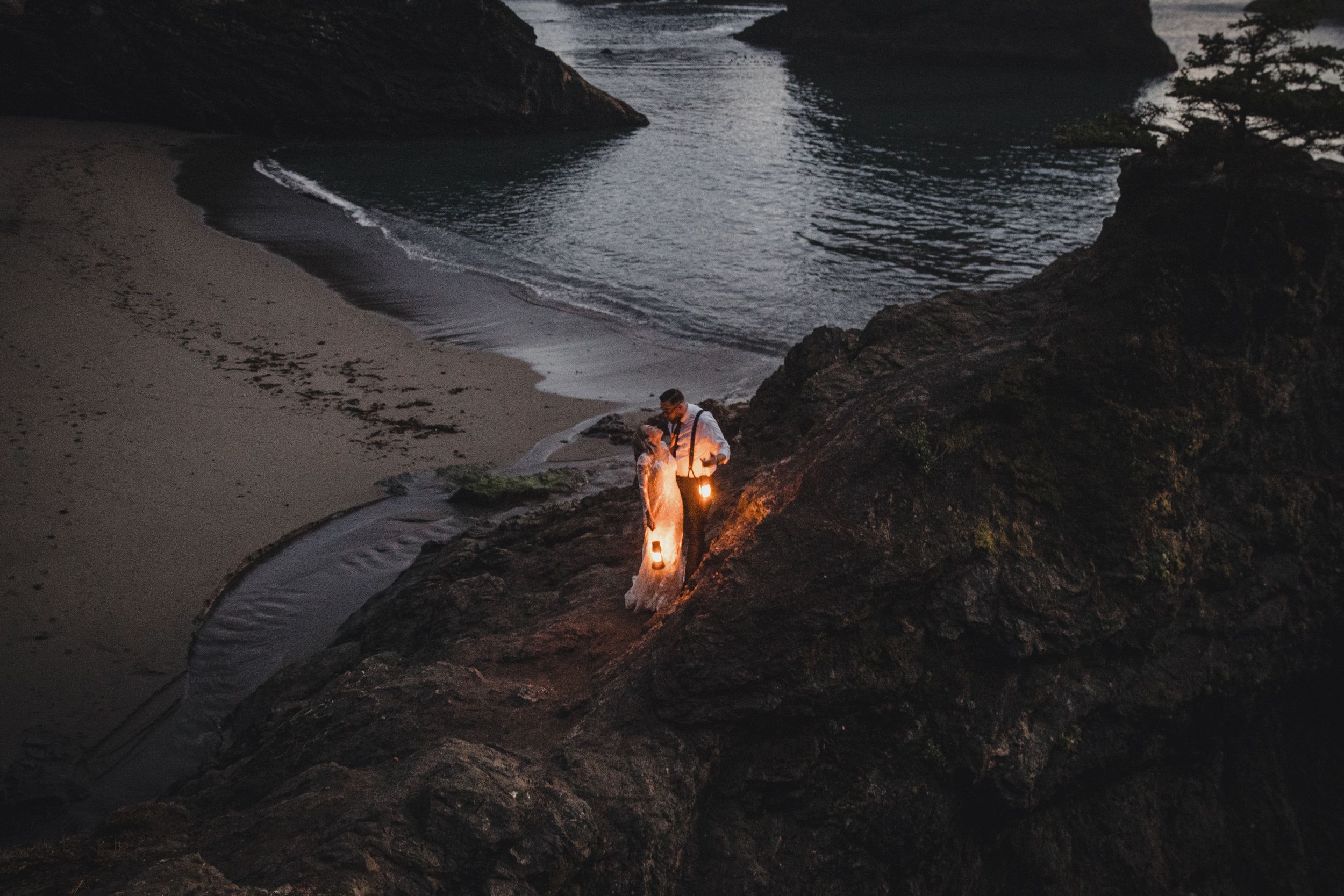 A glowing campfire illuminates a secluded beach cove at dusk with dark cliffs and gentle waves in the background.
