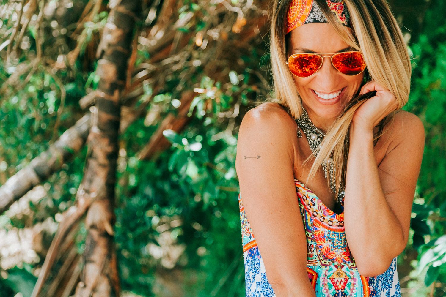 Woman in floral dress and sunglasses posing playfully against lush green background.