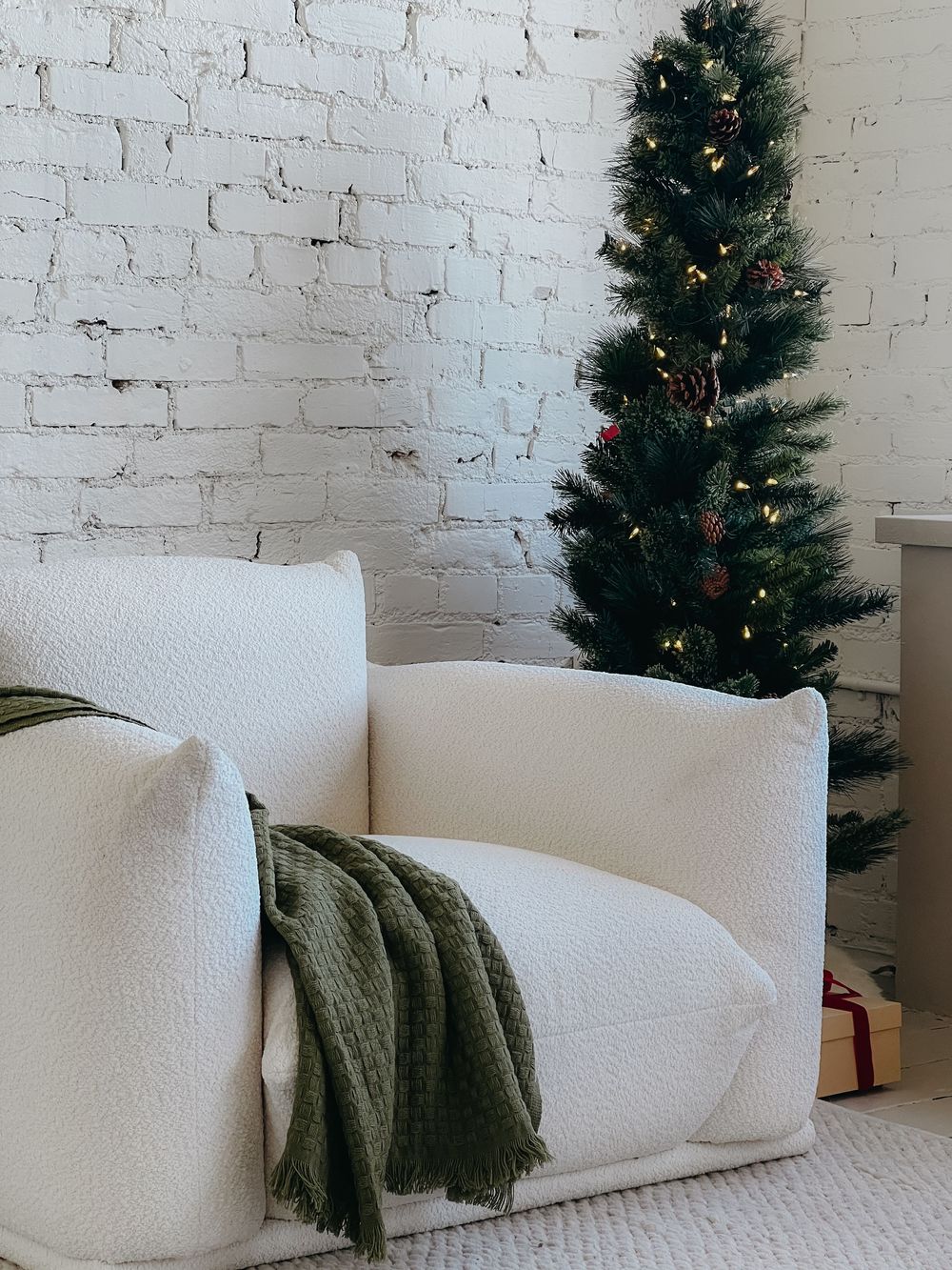 White curved modern sofa with green throw blanket next to decorated Christmas tree against white brick wall.