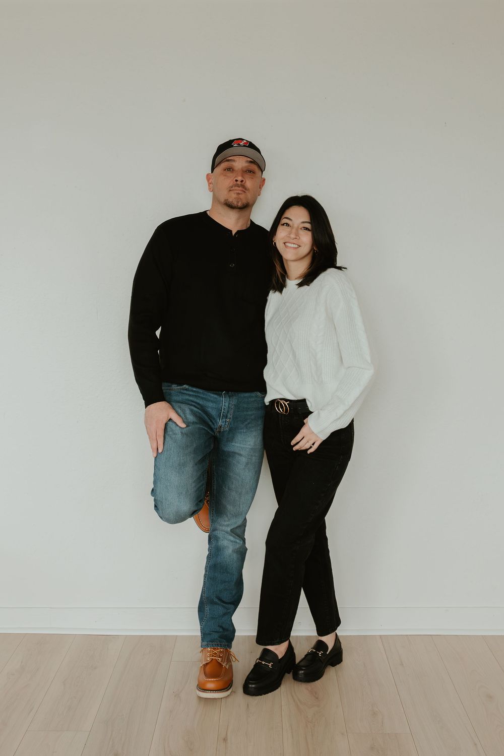 A couple poses together against a white wall in matching black and white outfits and casual denim.