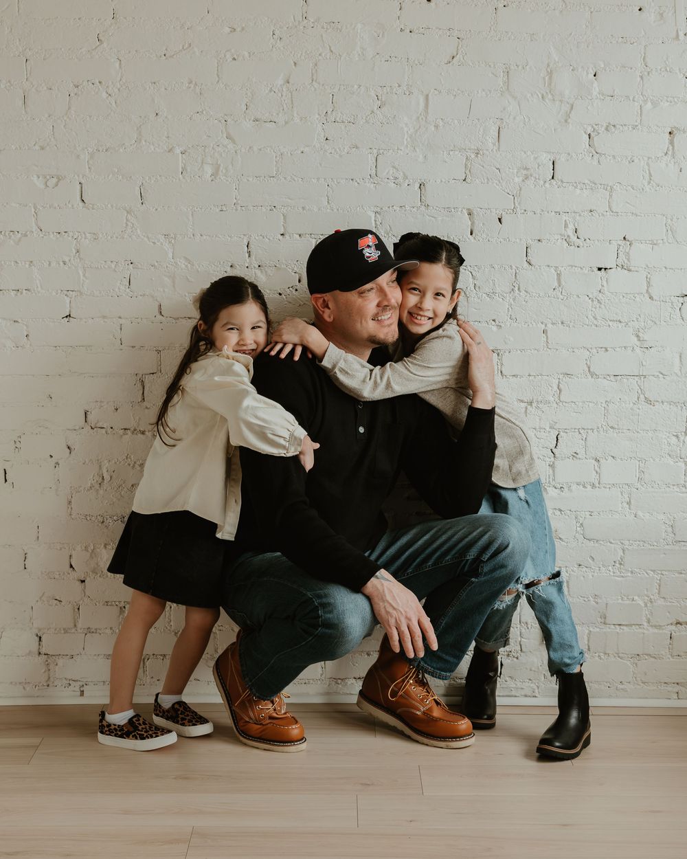 A family in coordinated black outfits sits together against a white brick wall in a minimalist studio setting.