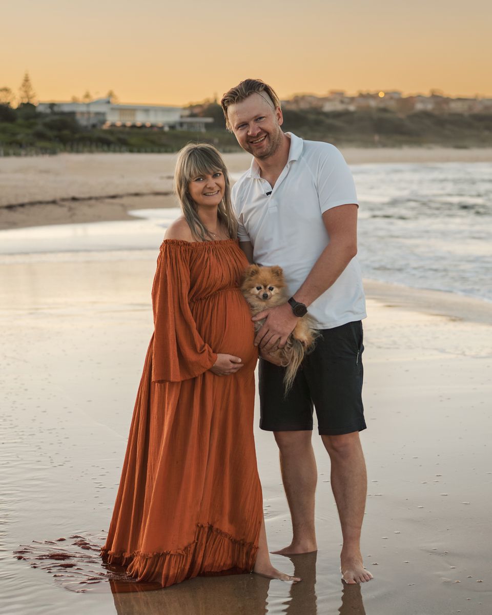 Couple with their small Pomeranian puppy enjoying a romantic sunset along a sandy beach during their maternity photoshoot in Sydney Maroubra