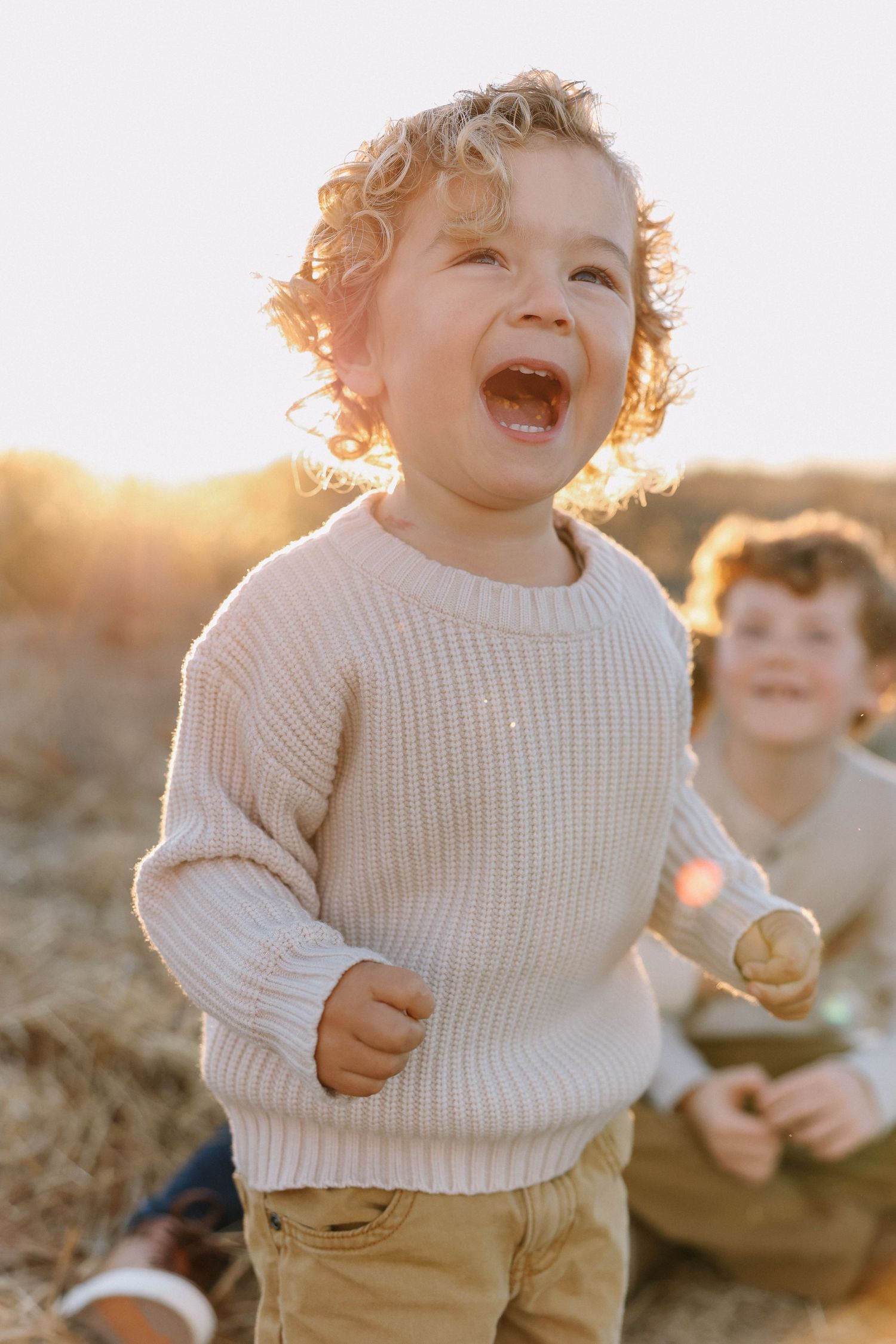 Toddler in white sweater laughs joyfully while playing outdoors during golden hour sunset.