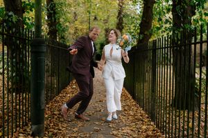 A couple walks along a tree-lined path with iron gates and fallen autumn leaves during a photo session.