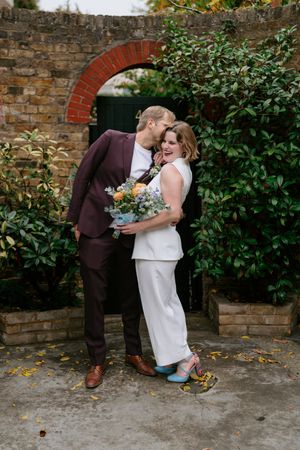 A couple shares a tender moment against a rustic brick wall in a garden setting during their wedding celebration.
