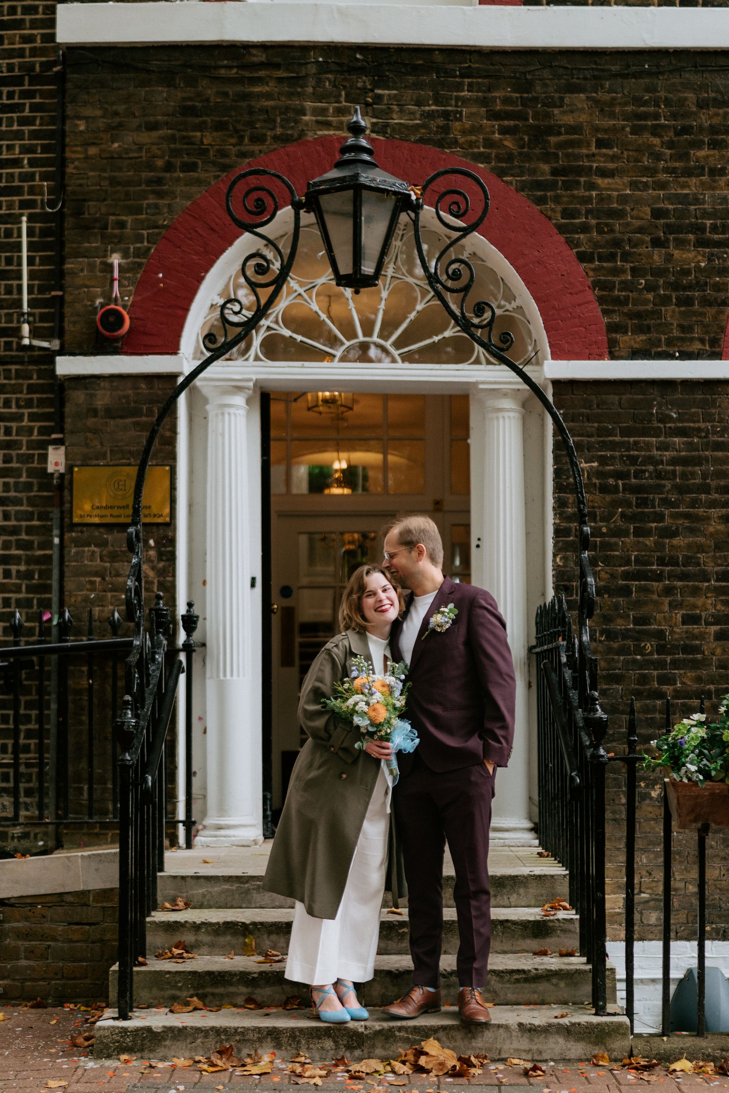 Couple shares intimate moment on brick building steps with elegant white columns and black iron railings.