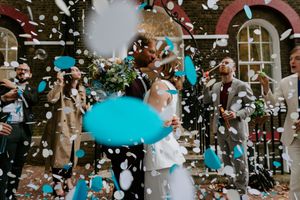 Confetti and blue balloons float through the air at an outdoor wedding celebration with white windows in background.