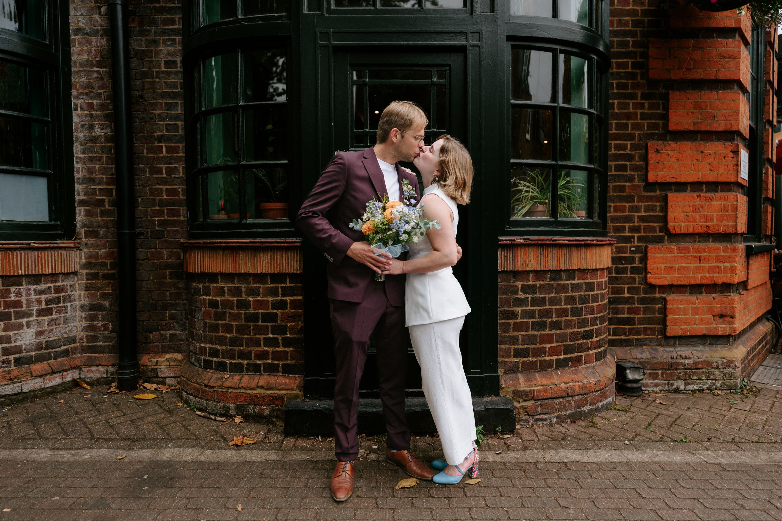A couple shares a romantic moment outside a historic brick building with a curved bay window.