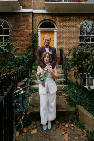 A couple stands on brick townhouse steps with autumn leaves and decorative ironwork in an urban setting.