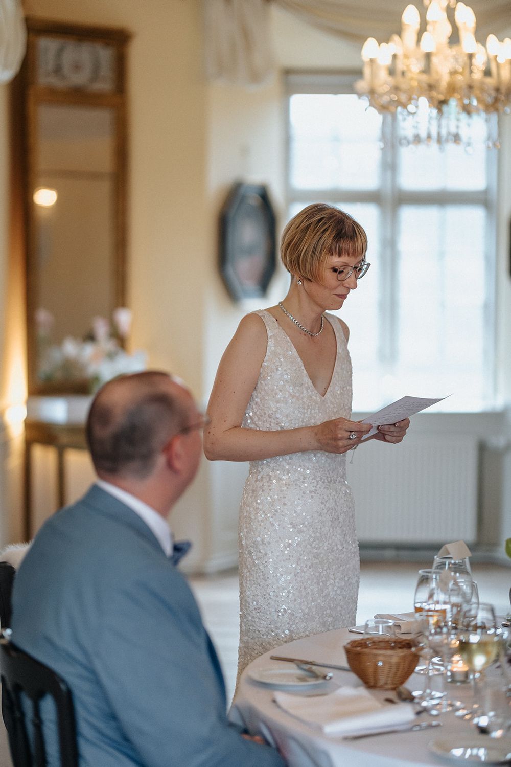 A wedding speech is delivered at an elegant reception with crystal chandeliers and formal table settings.