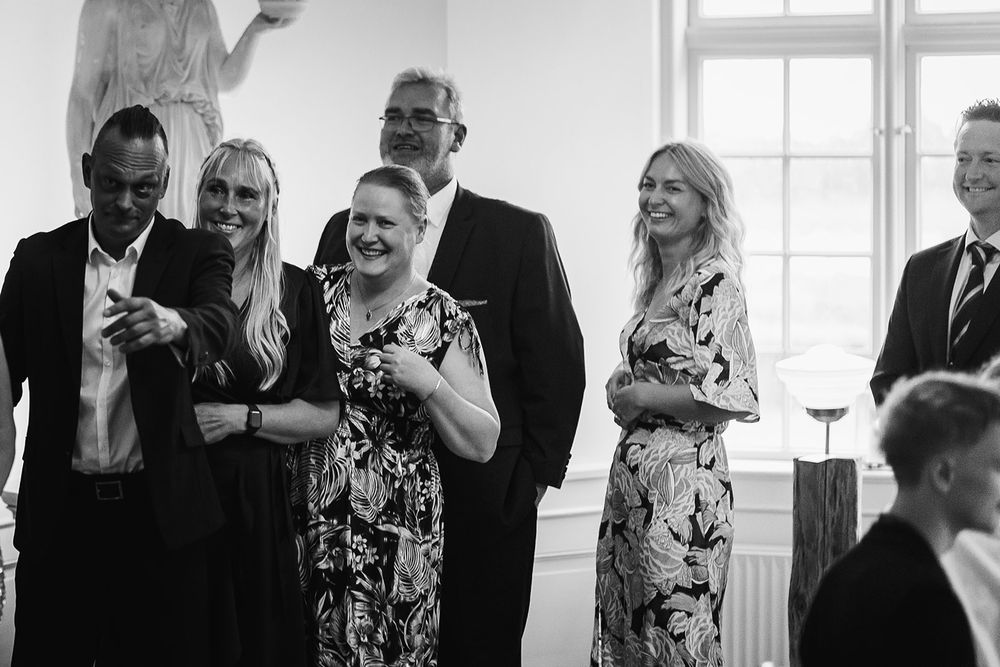 A group of formally dressed guests mingle and socialize at an elegant indoor event with large windows.
