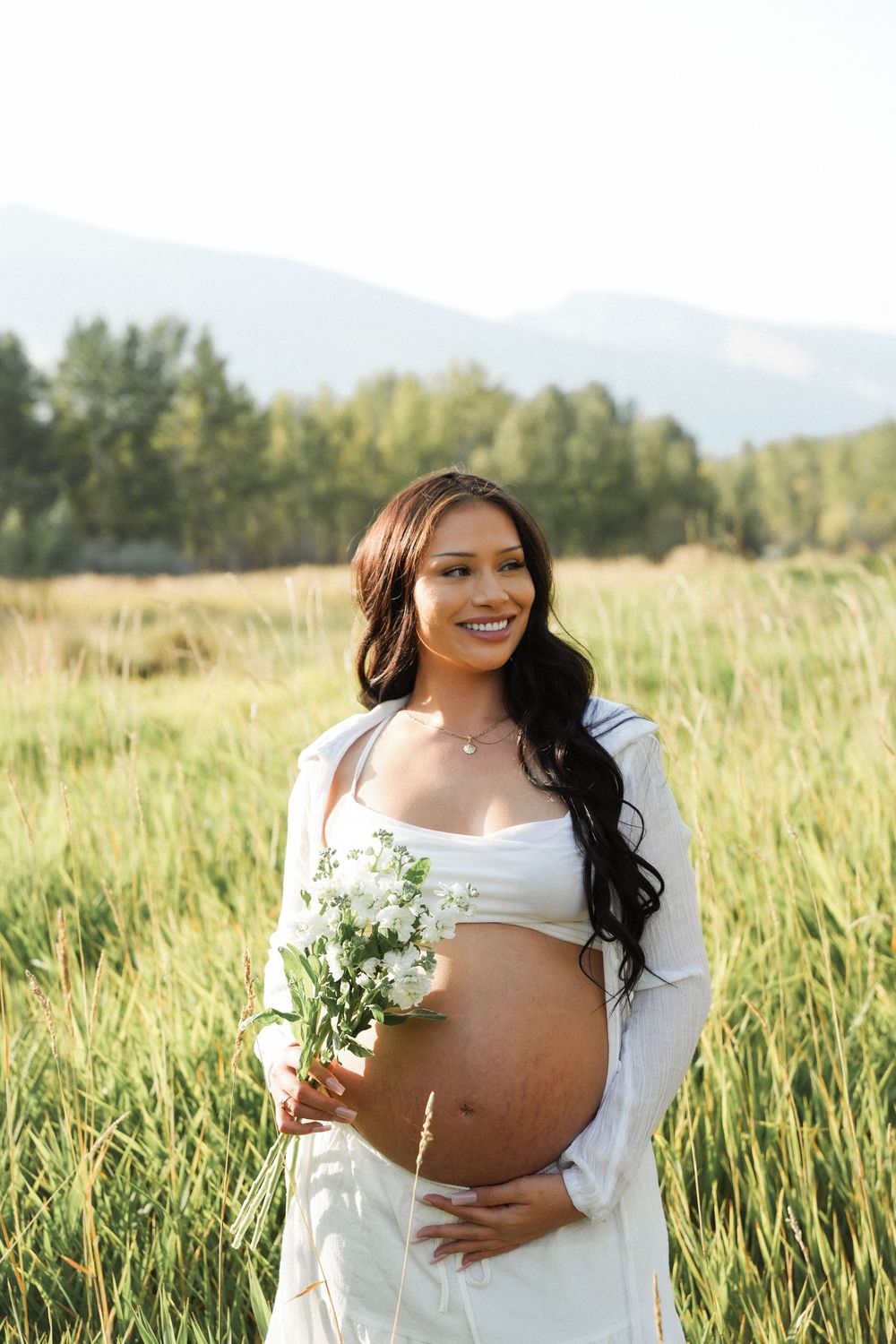 A pregnant woman in a white dress holds wildflowers while standing in a sunlit meadow with mountains in the background.