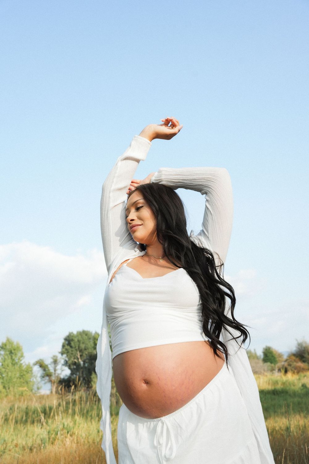 Pregnant woman in white flowing dress stretches gracefully in a sunlit meadow against blue sky.