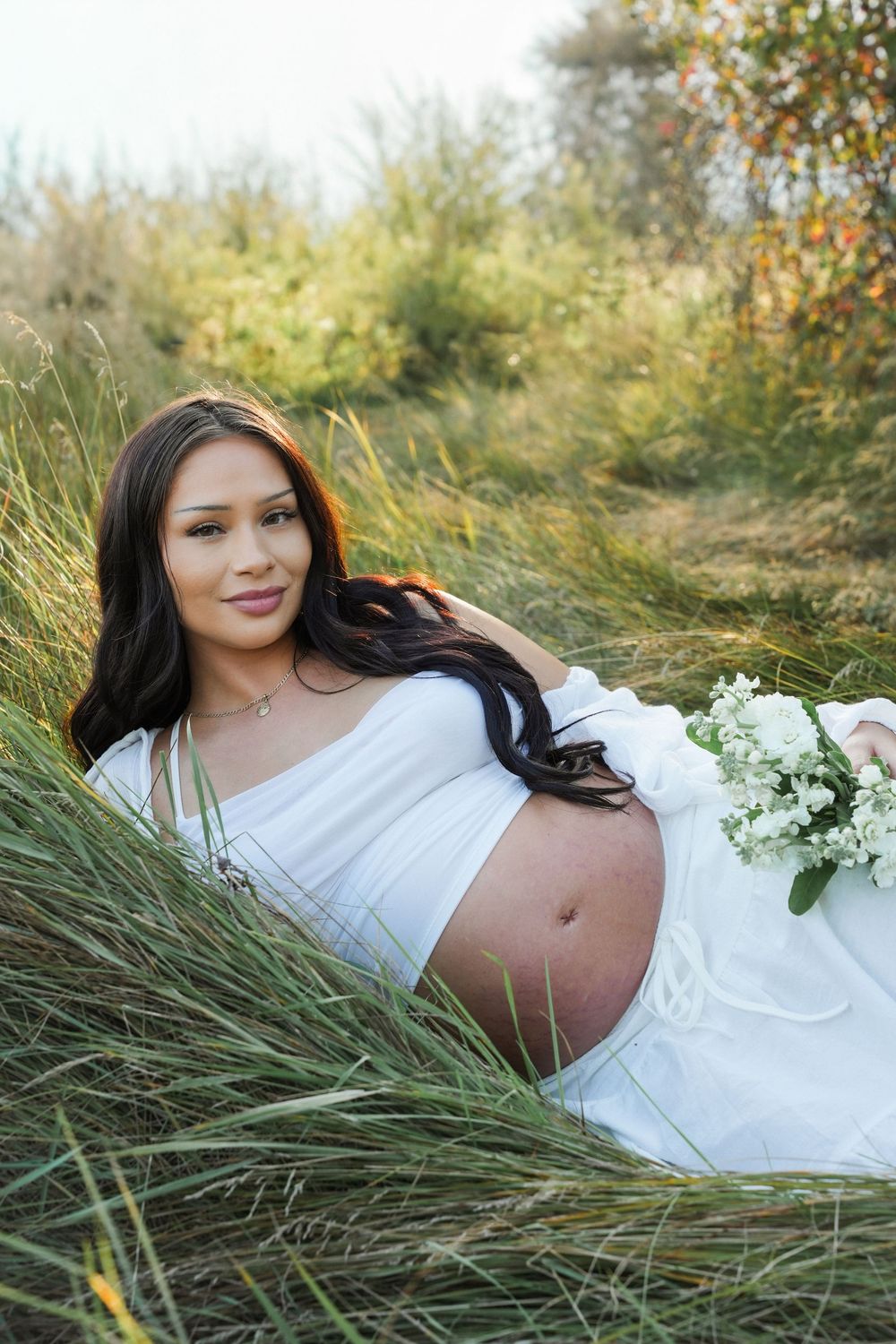 A pregnant woman in a white dress lies gracefully in tall grass during an outdoor maternity photoshoot.