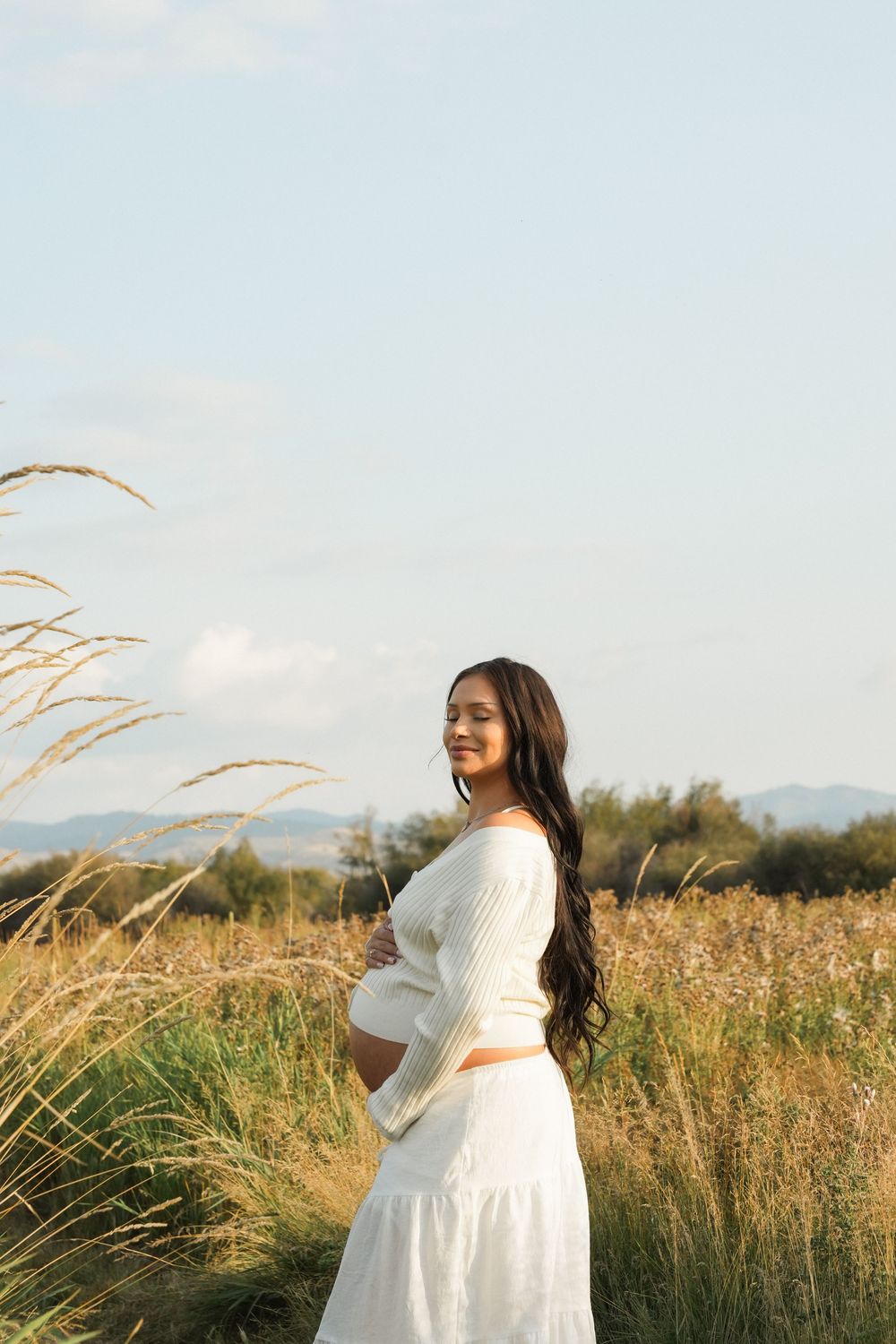 Expectant woman in white dress stands in golden field with tall grass swaying in breeze at sunset.