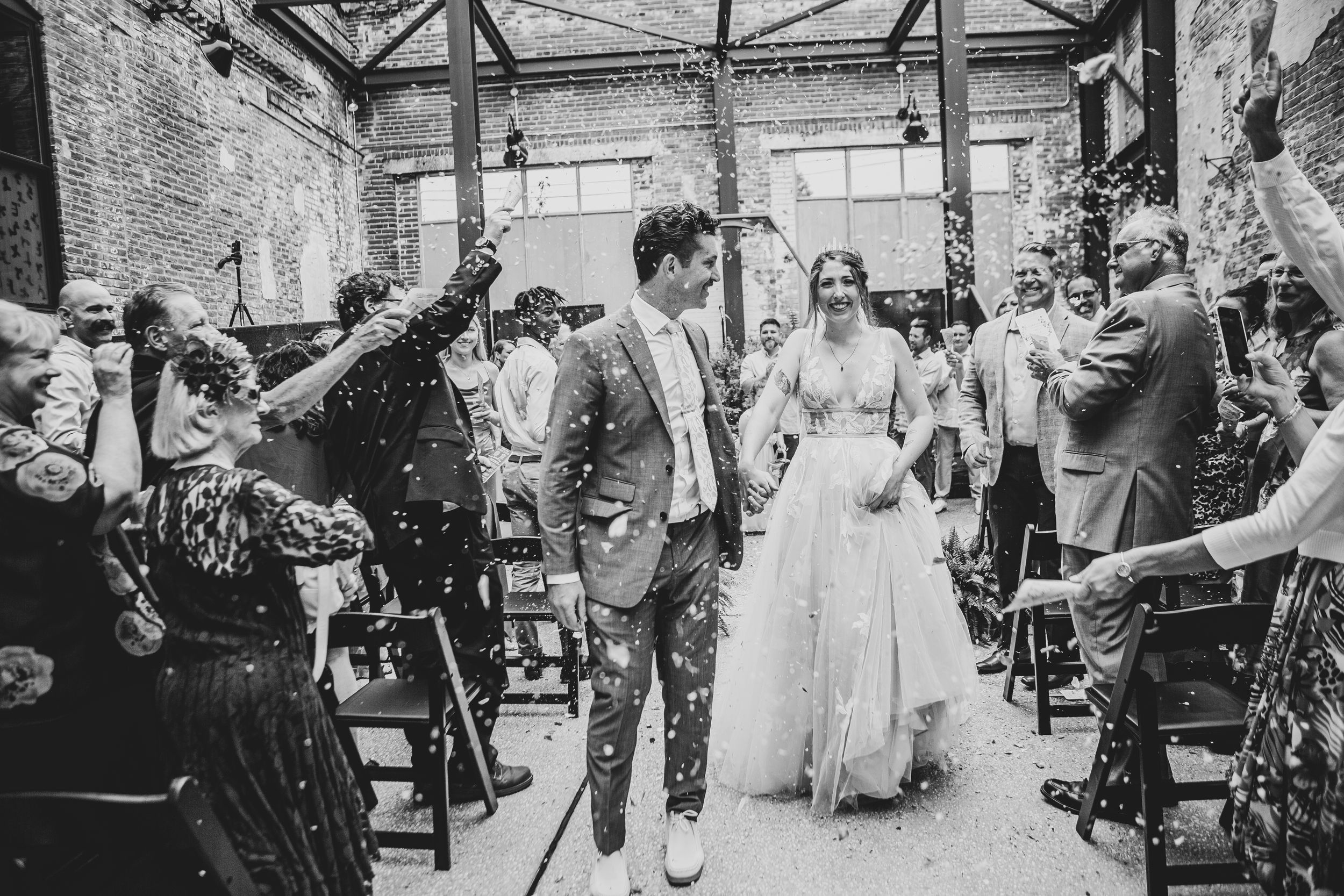 A black and white photo of a wedding celebration in an industrial venue with guests throwing confetti.