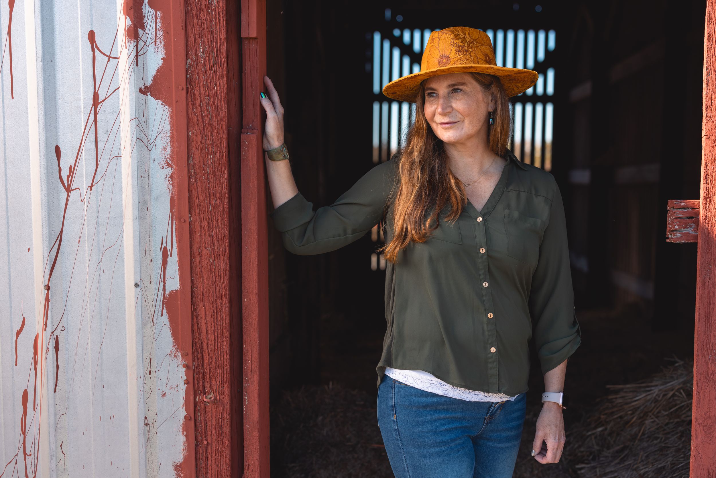 A woman in a brown hat and olive shirt poses against a rustic red barn door.