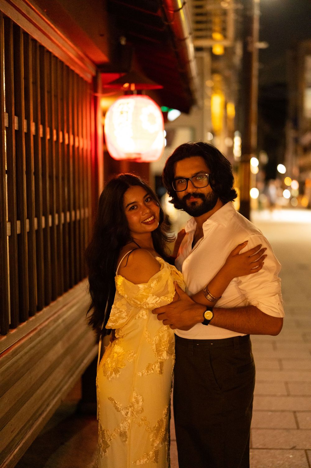Smiling couple embracing on a warmly lit street at night near a red Japanese lantern.