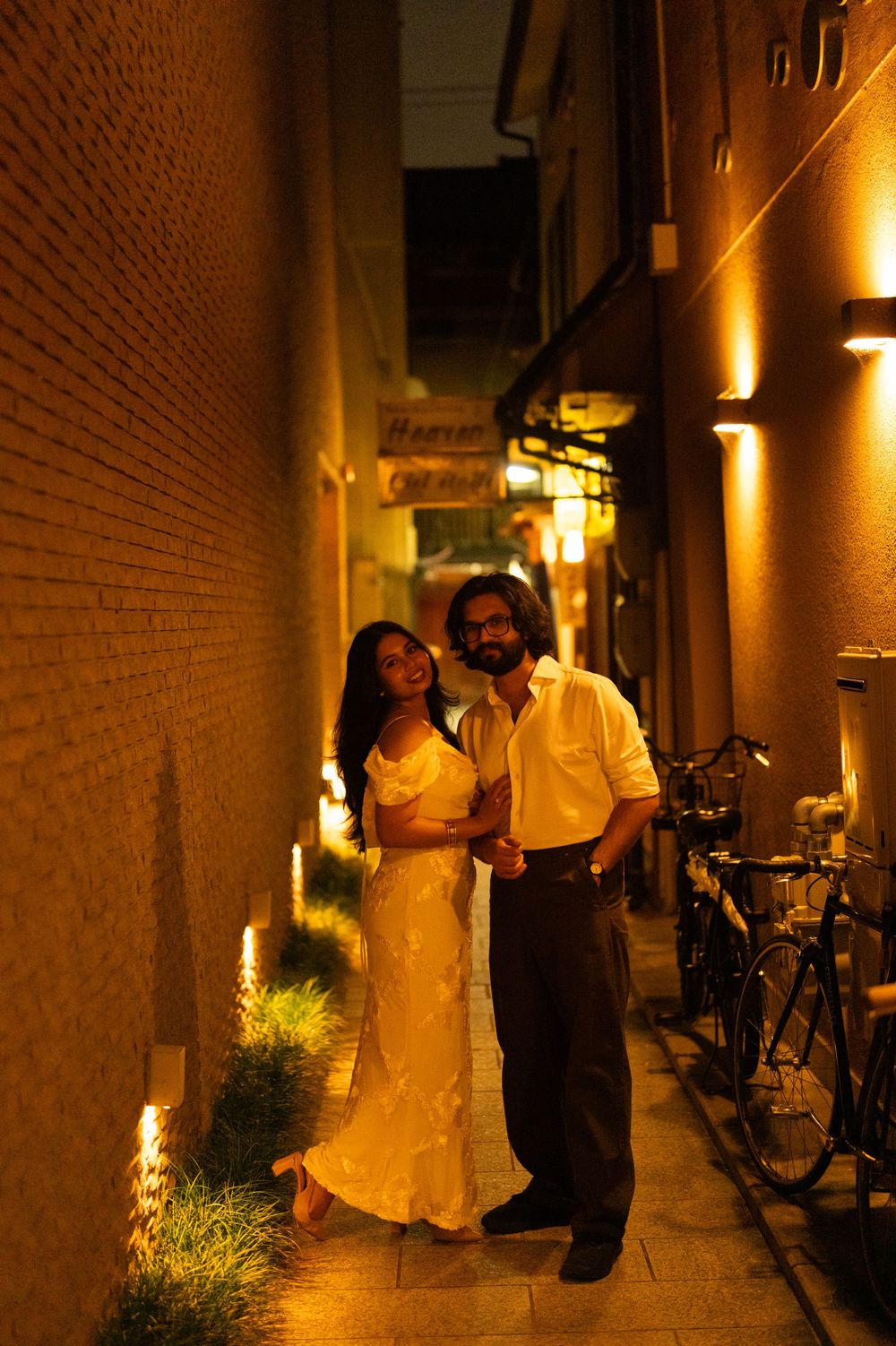 Couple posing together in a warmly lit narrow alley at night with brick walls and bicycles.