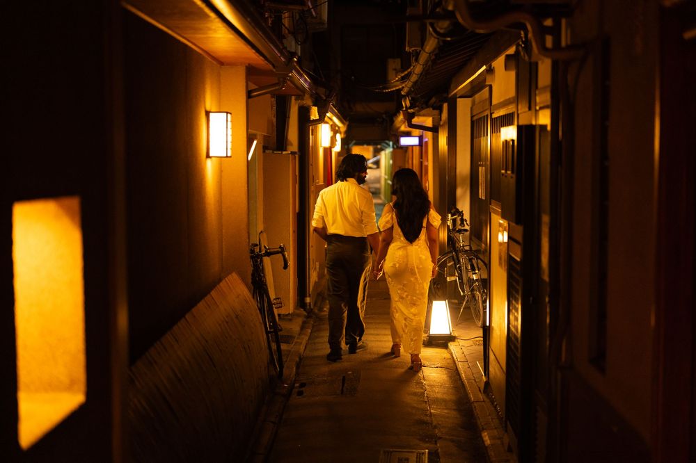 Couple walking hand in hand through a narrow lantern-lit Japanese alleyway at night.