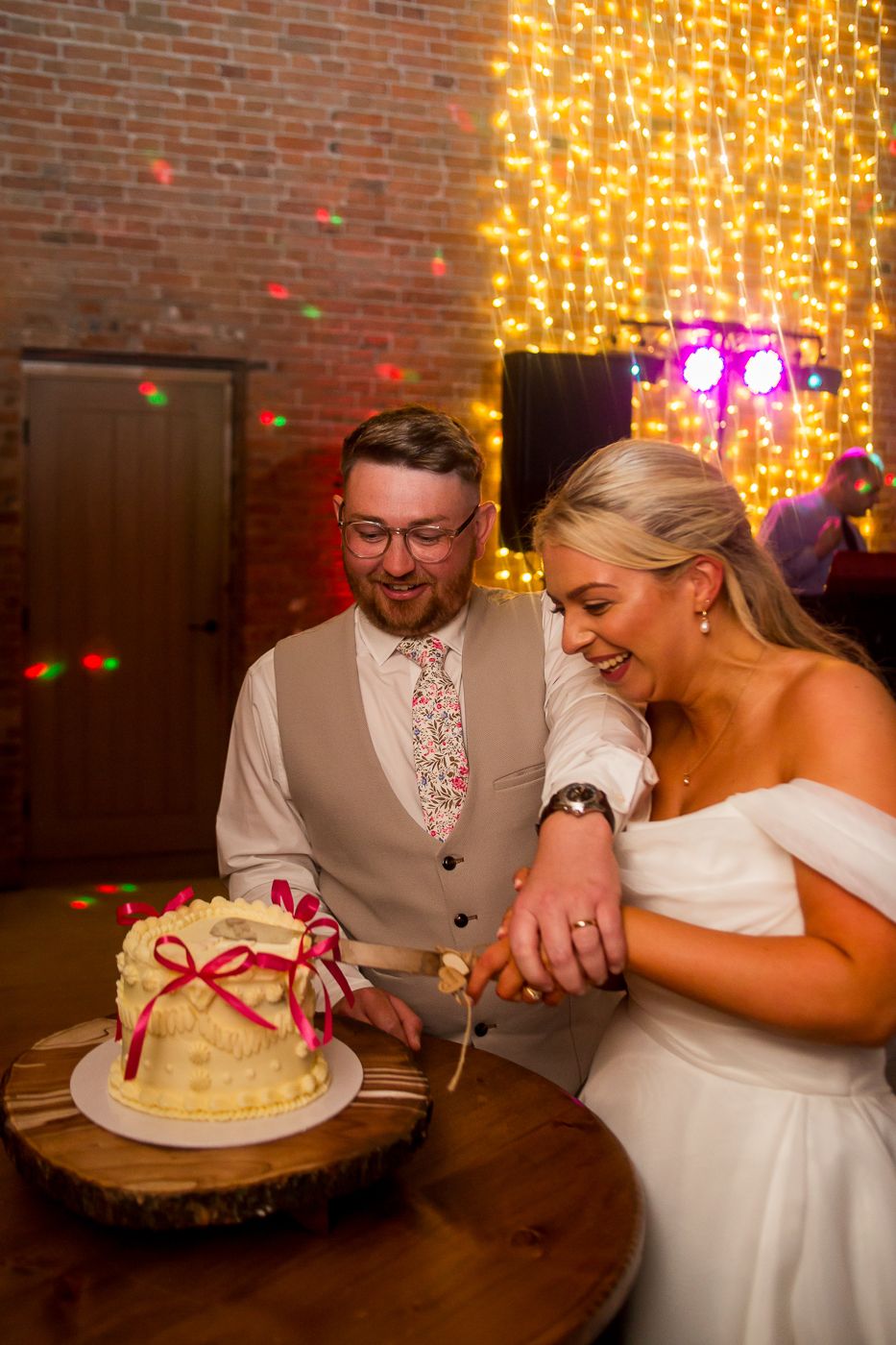 A couple cuts into their wedding cake decorated with a red bow on a wooden table with twinkle lights in the background.
