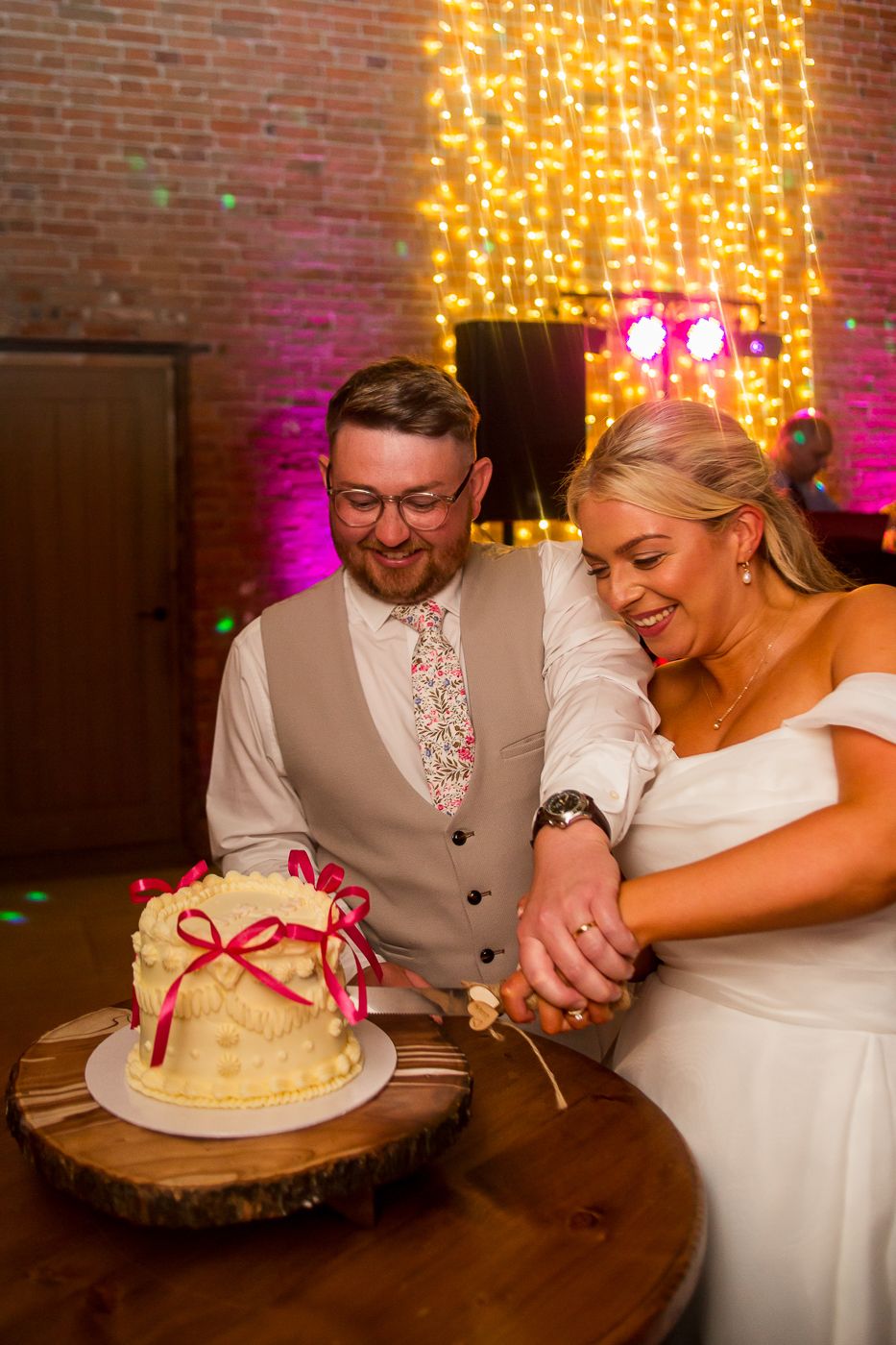 Newlyweds share a playful moment while cutting their white wedding cake with red ribbon accents.