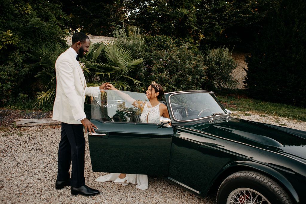 Groom opening the door of a vintage dark green Triumph convertible for his bride at a French estate.