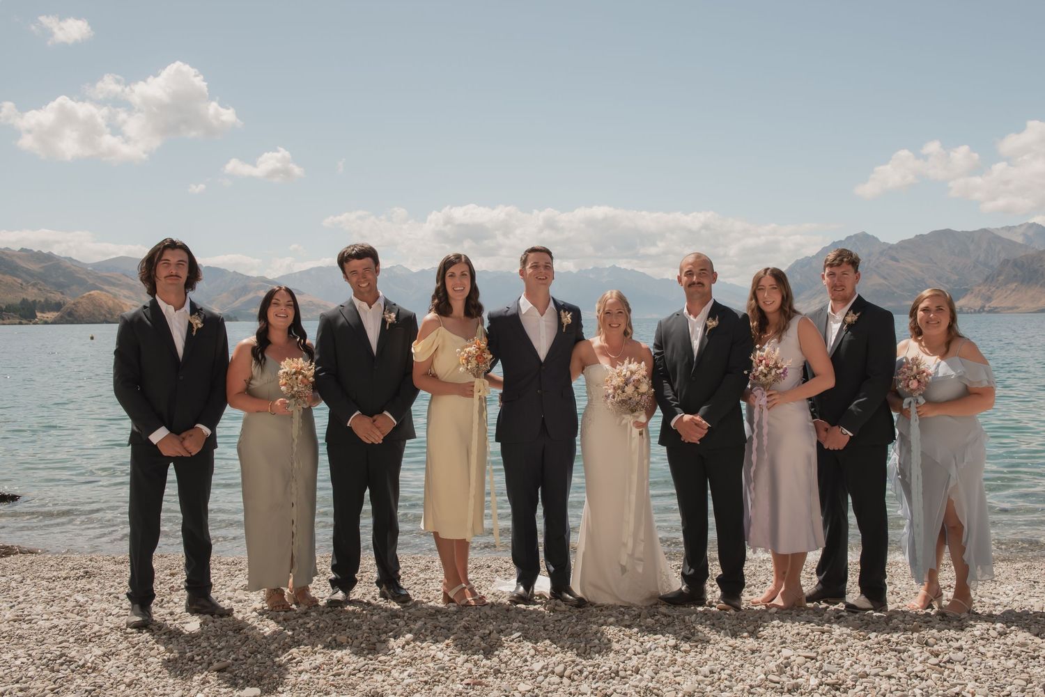 Wedding party members in formal attire stand together on a scenic beach with mountains in the background.