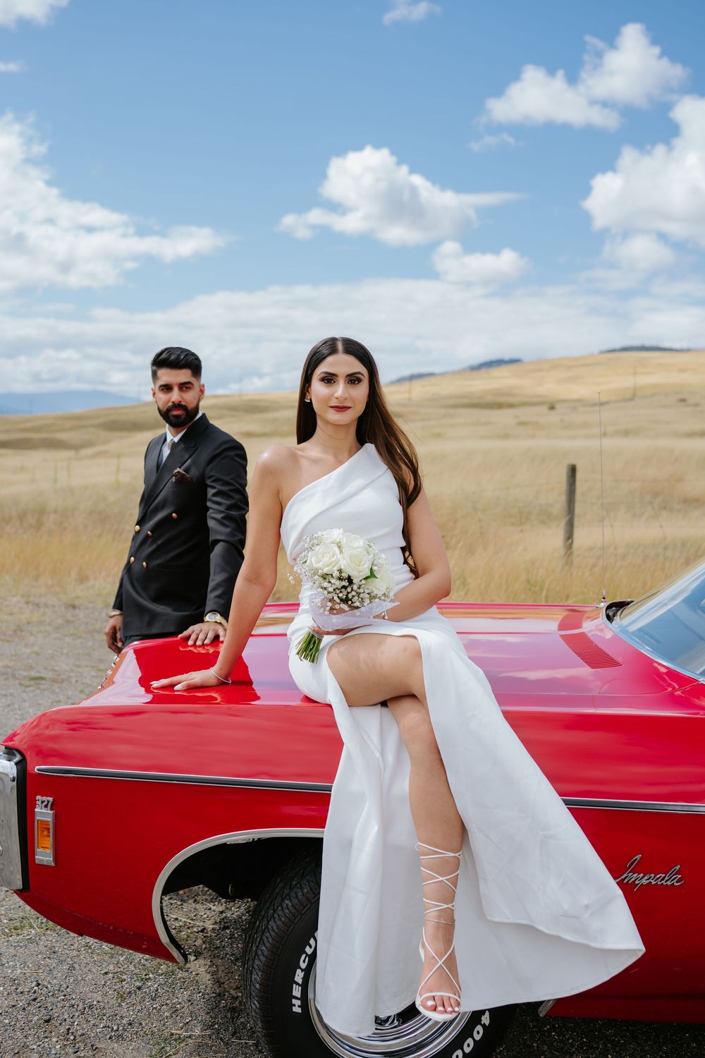 A couple poses with a classic red convertible car in a scenic field with rolling hills and blue sky.