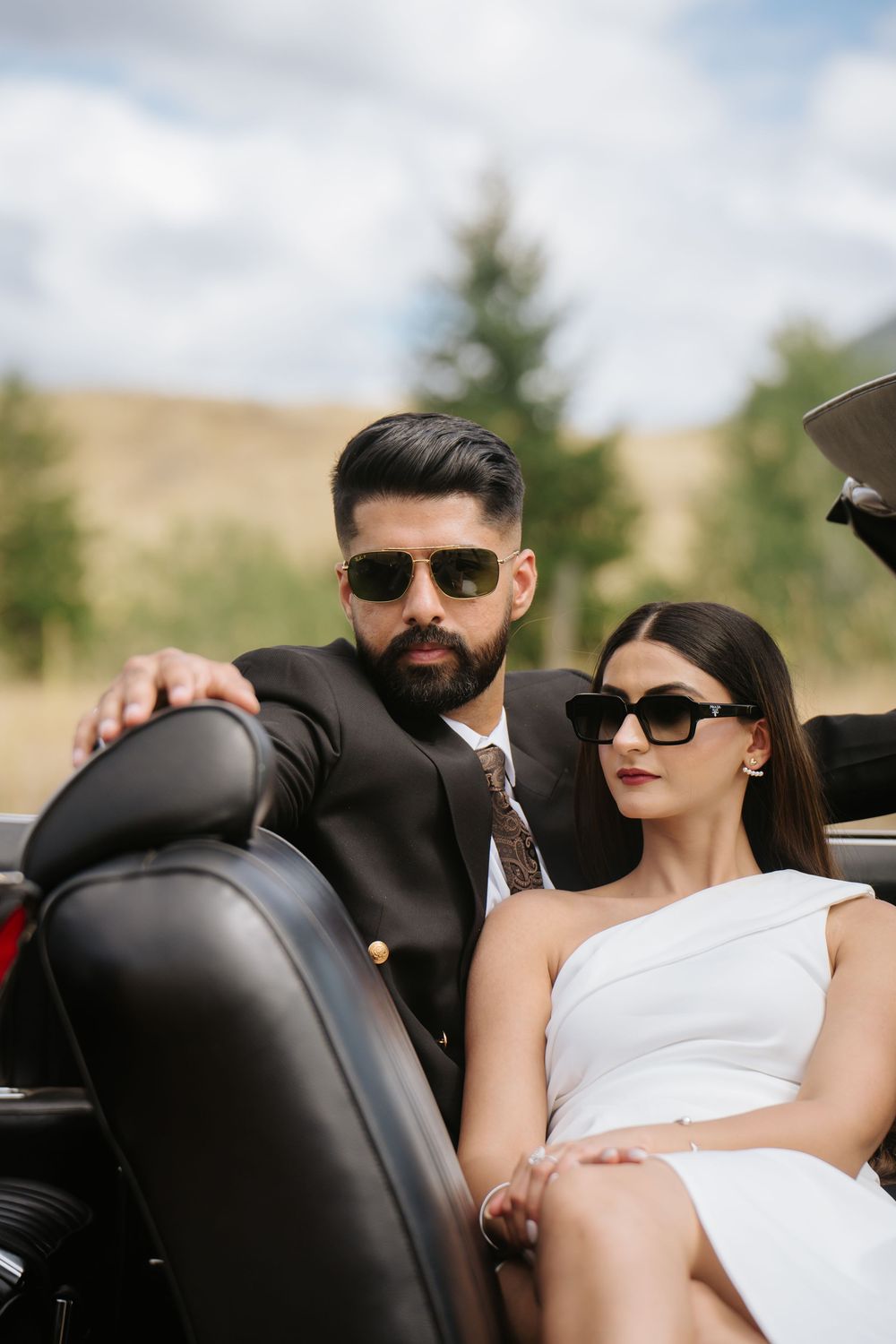 A stylish couple wearing sunglasses relaxes in the black leather seats of a convertible car.