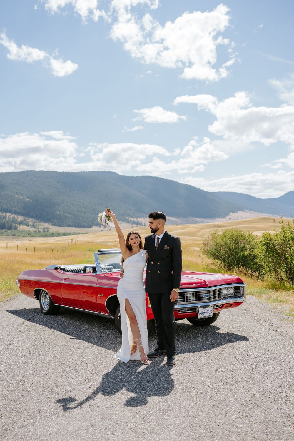 A couple poses with a classic red convertible car against a scenic mountain landscape on a sunny day.