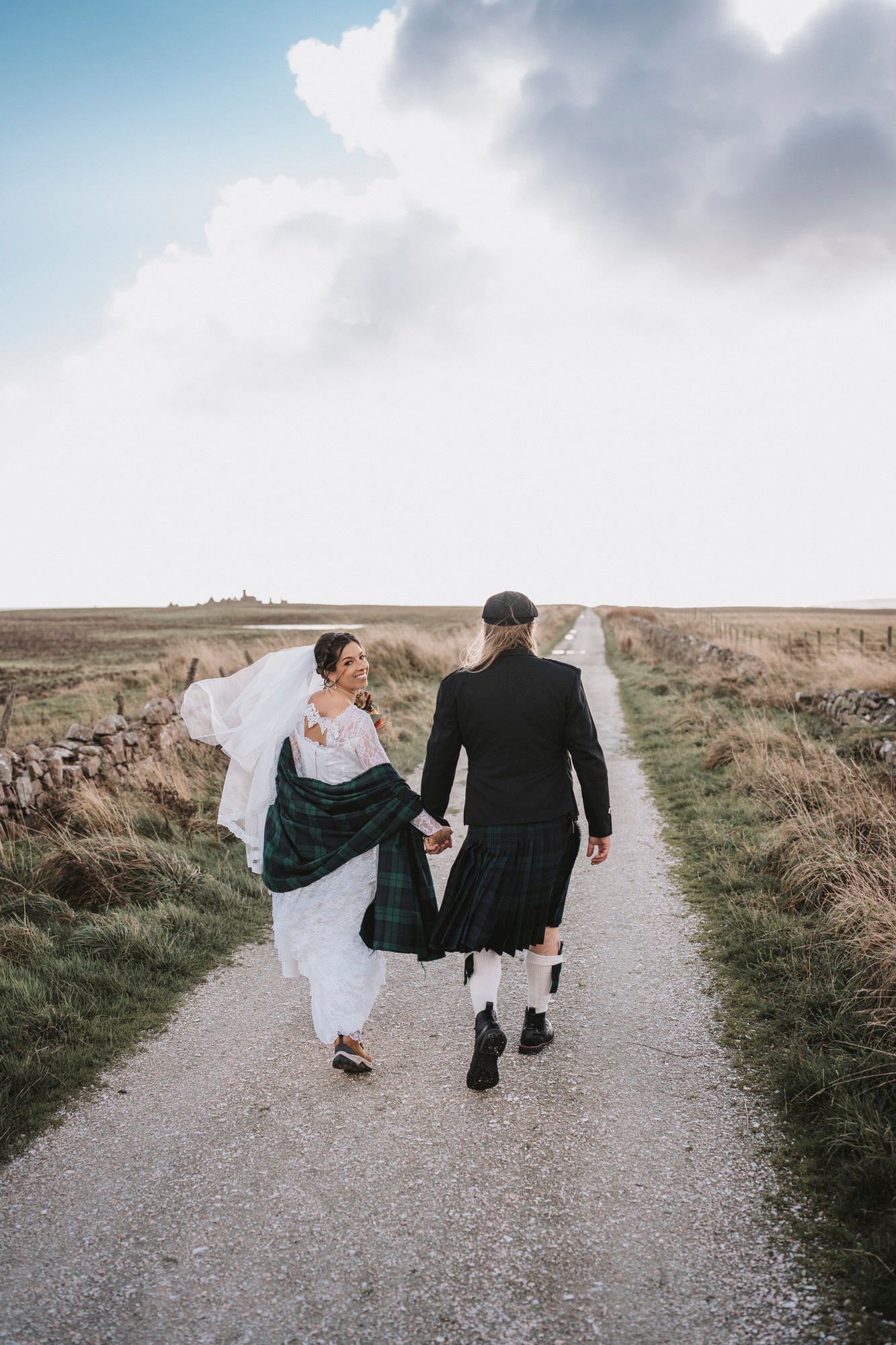 A couple in wedding attire walks along a scenic gravel path on a cloudy day in a rural landscape.