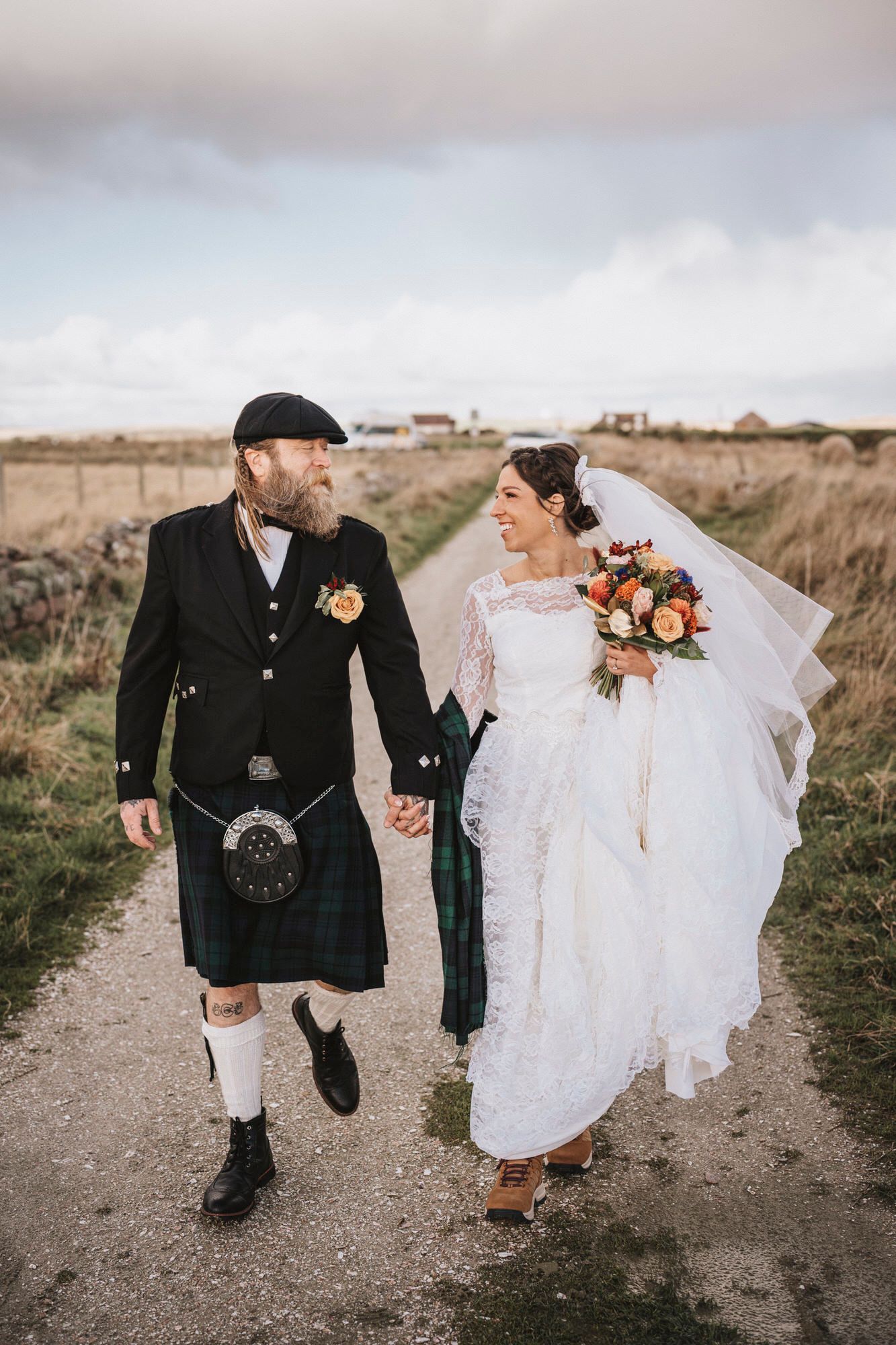A couple in Scottish wedding attire walking together on a dirt path through countryside fields.