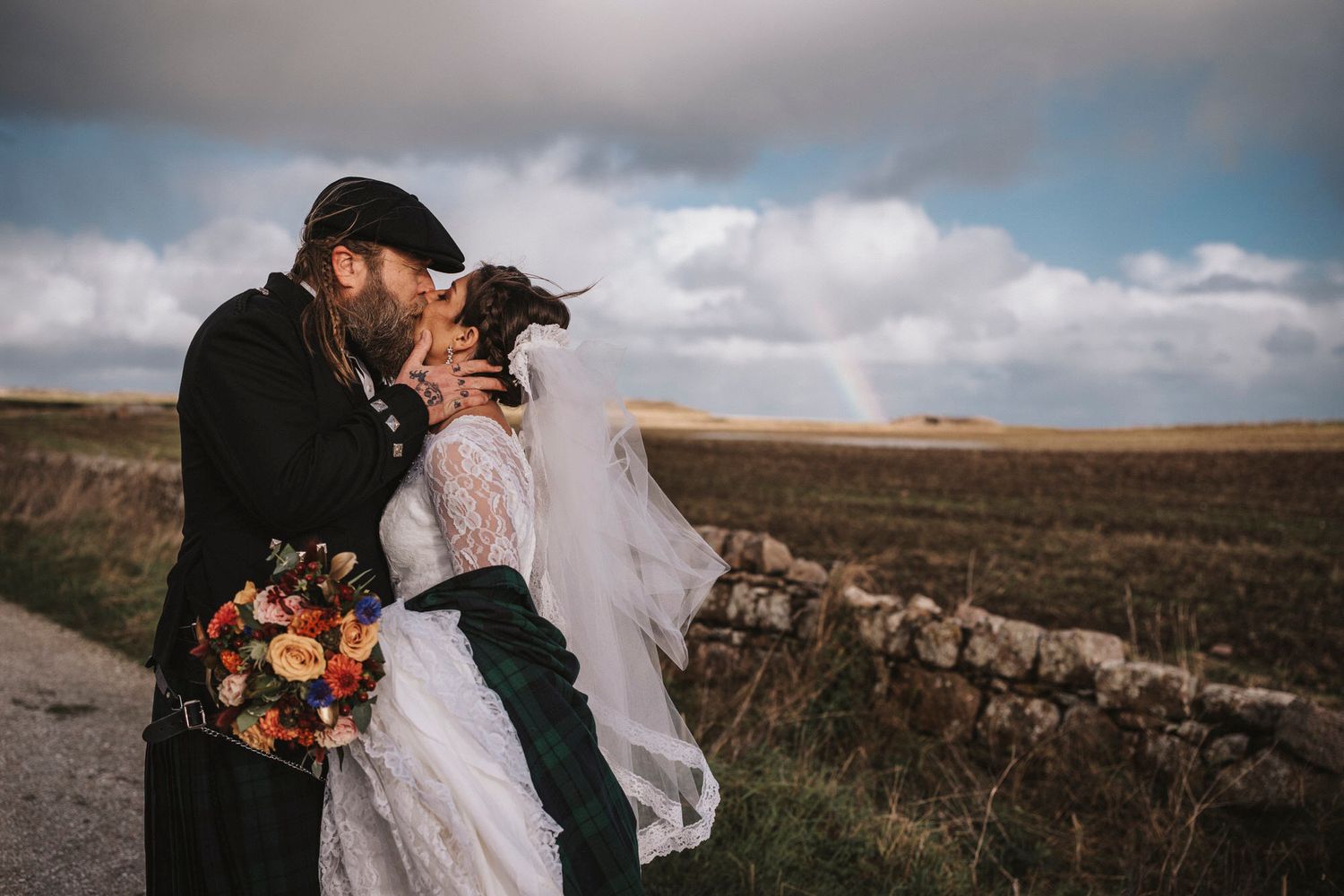 A couple embraces in a romantic moment against a cloudy rural landscape with stone walls and fields.