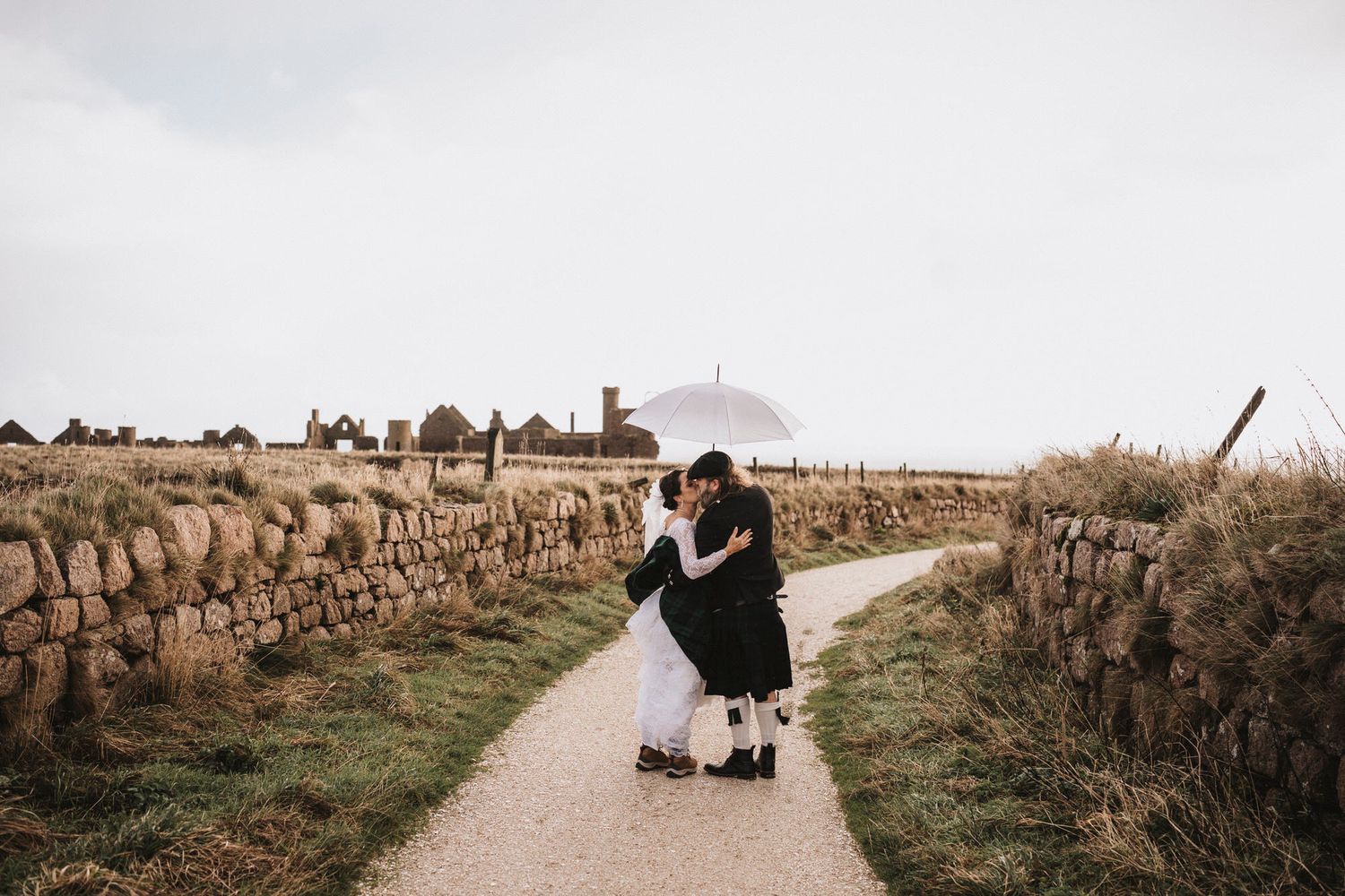 A couple in wedding attire walks along a rustic stone wall path through scenic countryside fields at sunset.